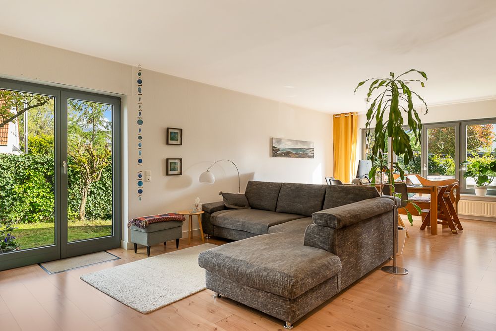 Helles Wohnzimmer mit grauem Ecksofa, Esstisch, großen Fenstern und Pflanzen.