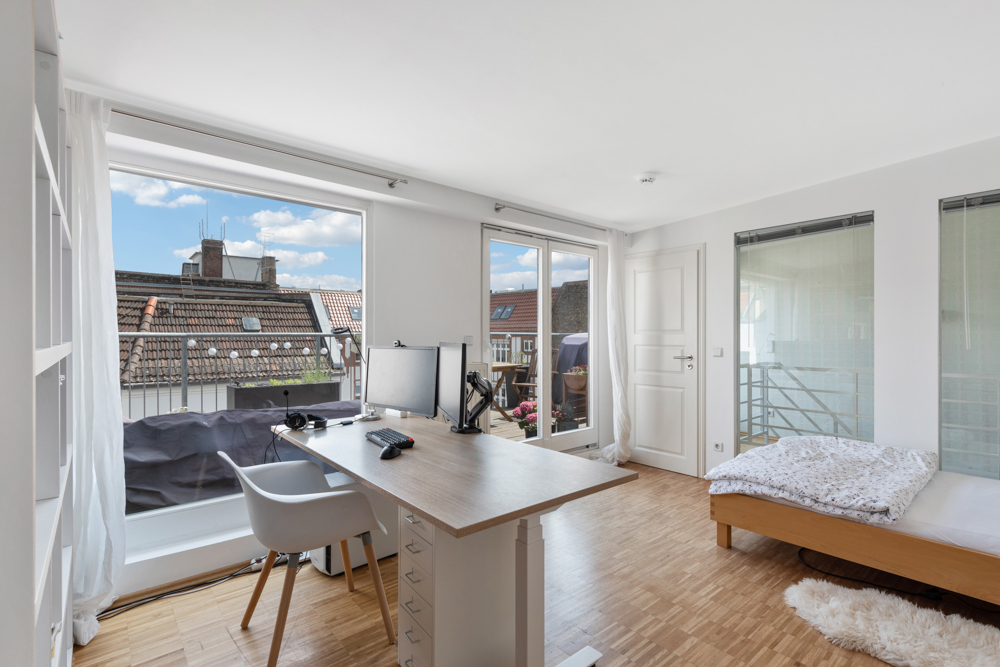 Helles Schlafzimmer mit Schreibtisch, Bett und Zugang zum Balkon, Holzboden und Dachfenster.