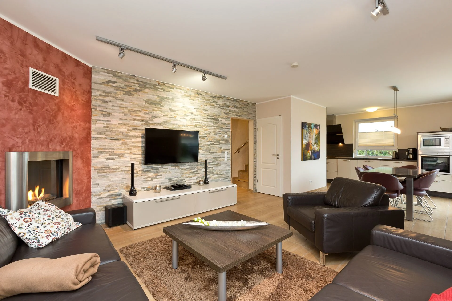 Modernes Wohnzimmer mit Kamin, Sitzgruppe, Fernseher und offener Küche im Hintergrund.