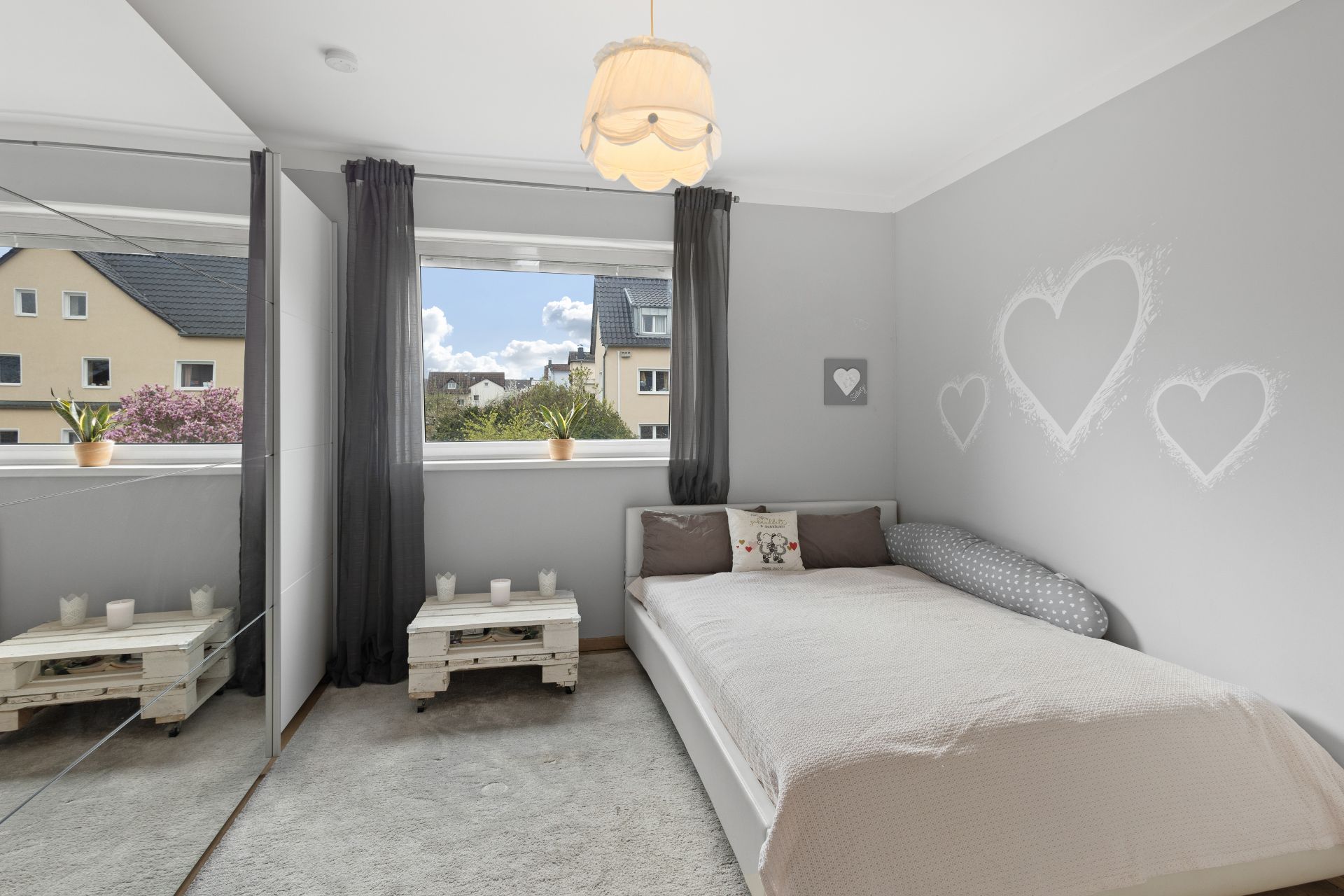 Helles Schlafzimmer mit großem Fenster, Bett, Nachttisch und Wanddeko in Herzform.