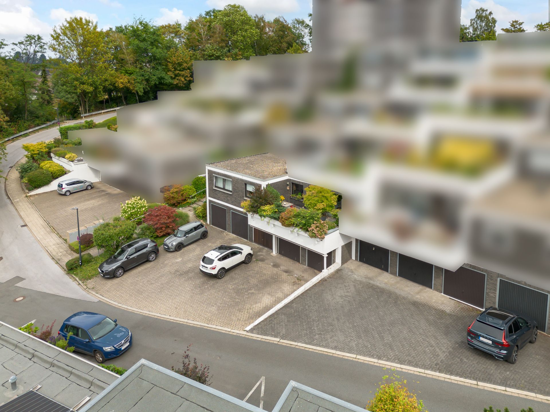 Mehrere Autos parken vor Garagen eines Wohngebäudes, umgeben von Bäumen und Gehwegen.