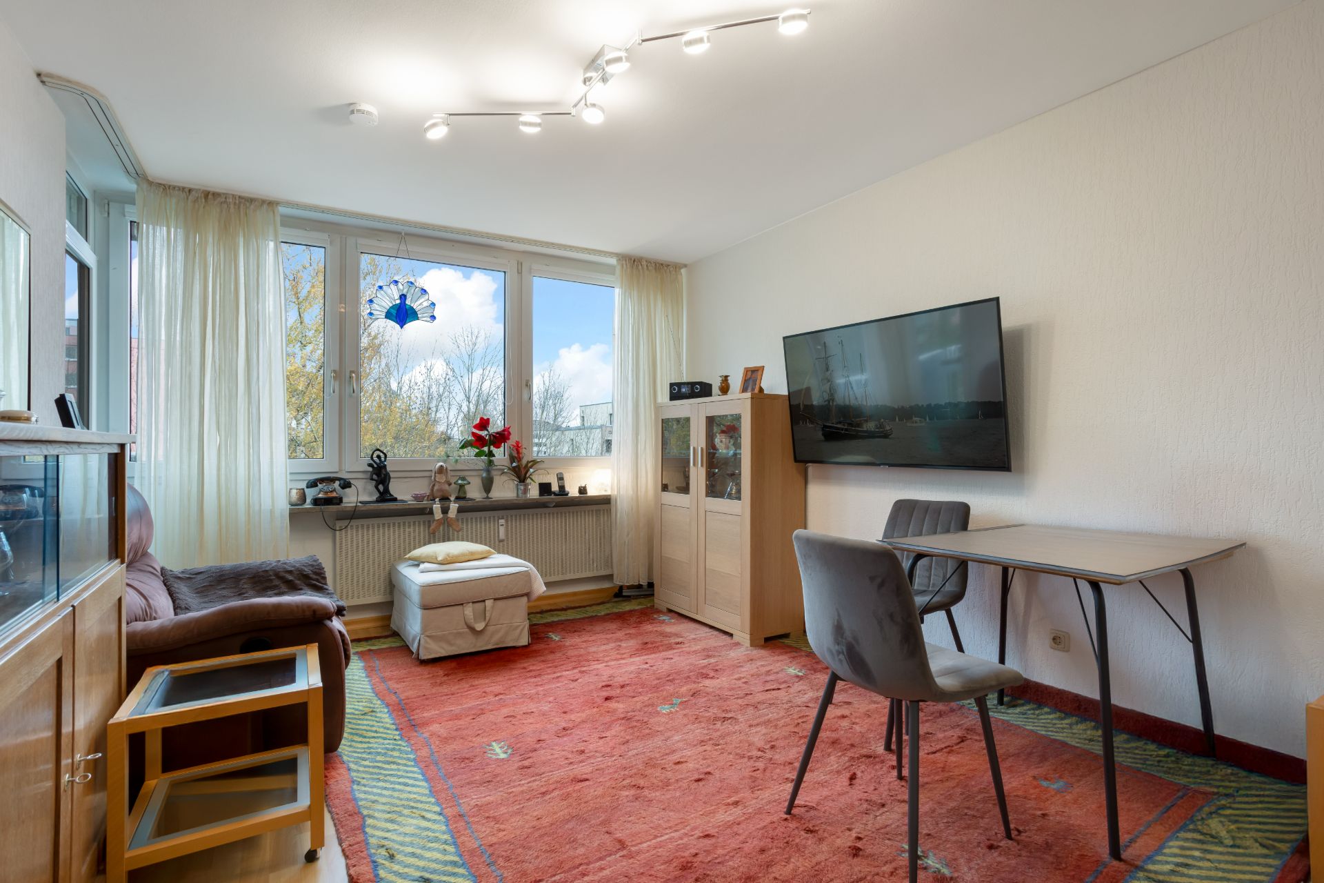 Wohnzimmer mit großem Fenster, rotem Teppich, Sofa, Tisch, Stühlen und Fernseher an der Wand.