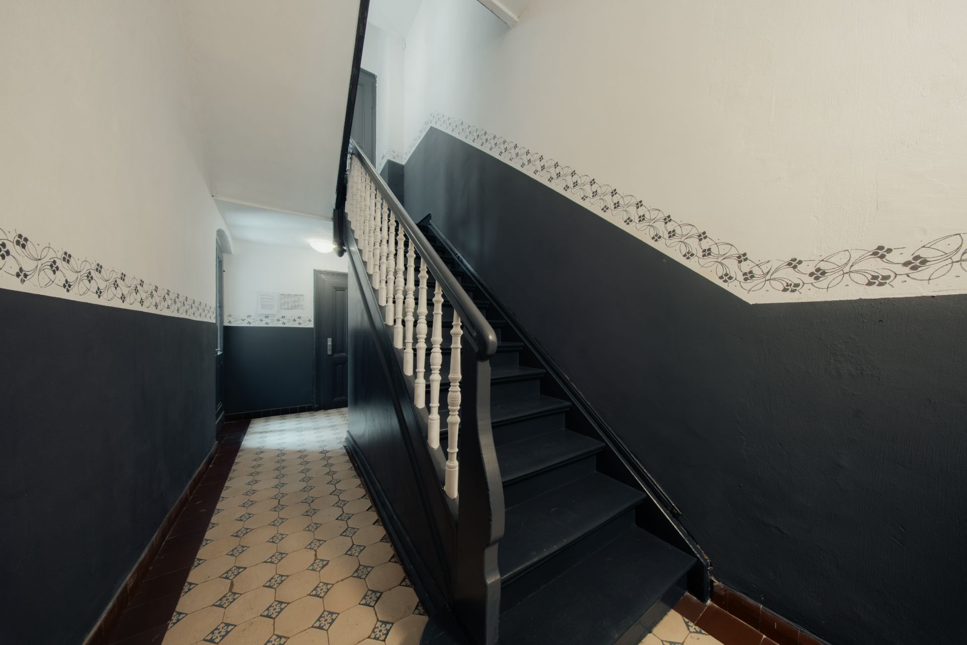 Treppenhaus mit dunkler Treppe und gemustertem Fliesenboden in einem Wohnhaus.