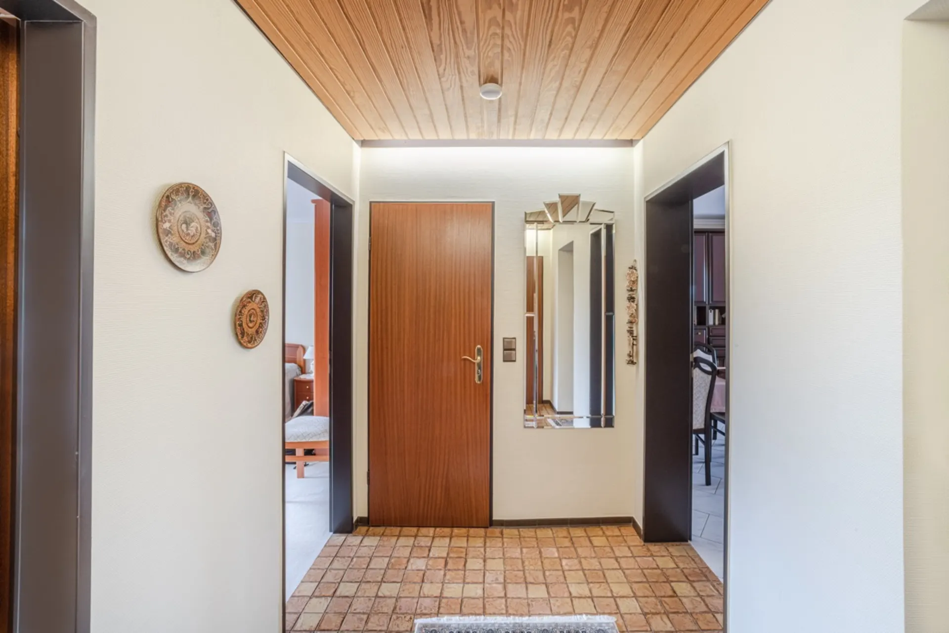 Flur mit Holztür, Spiegel, zwei Wandtellern und Fliesenboden.