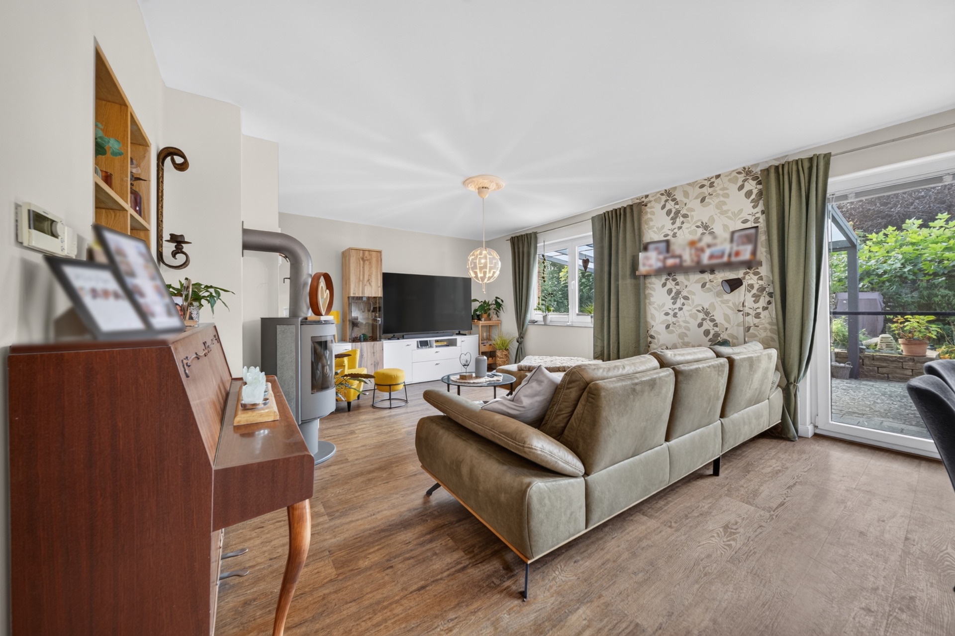 Helles Wohnzimmer mit grünem Sofa, TV, Holzfußboden und Blick auf den Garten durch große Fenster.