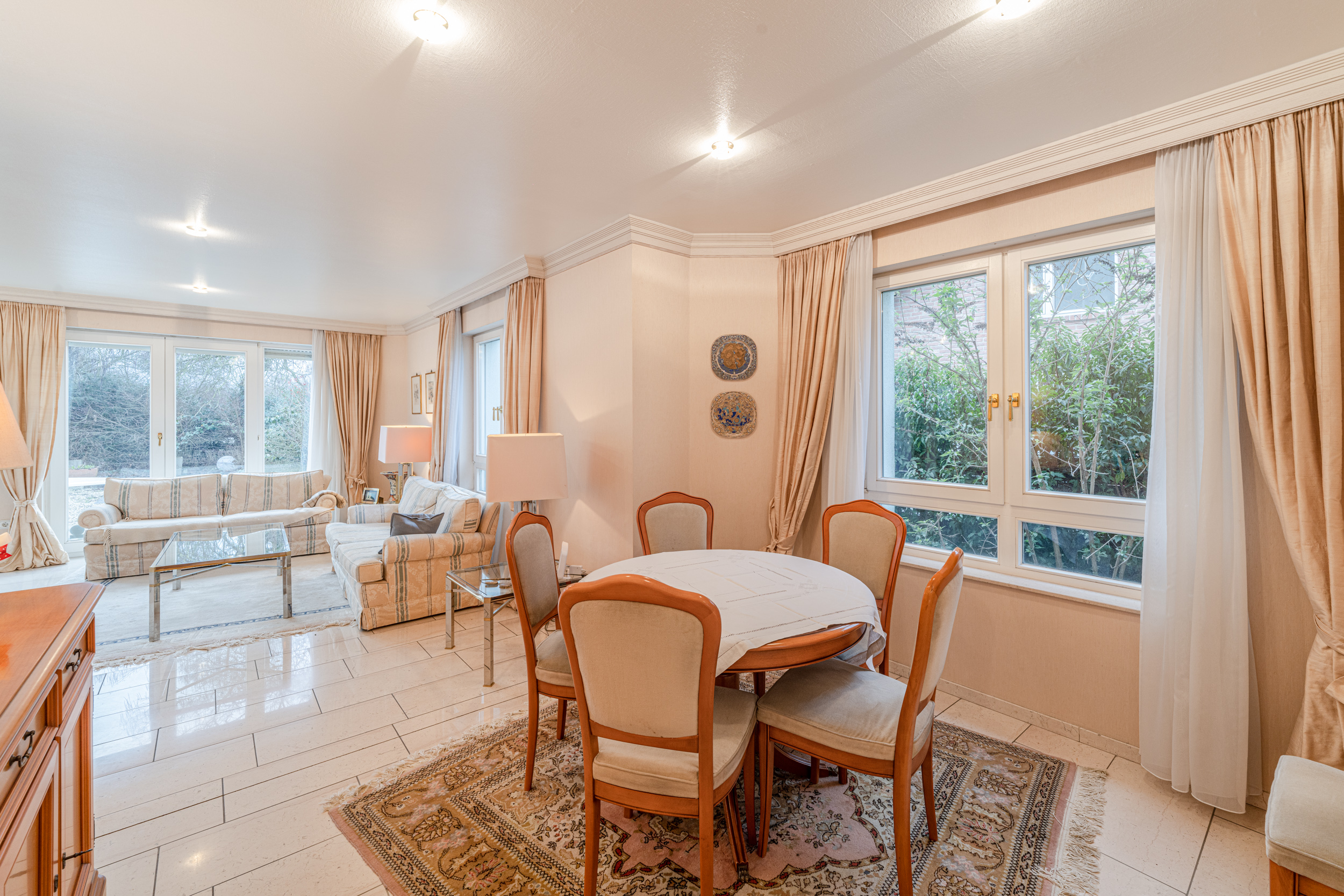 Helles Wohnzimmer mit Esstisch, Stühlen, Sofas, großen Fenstern und Vorhängen.