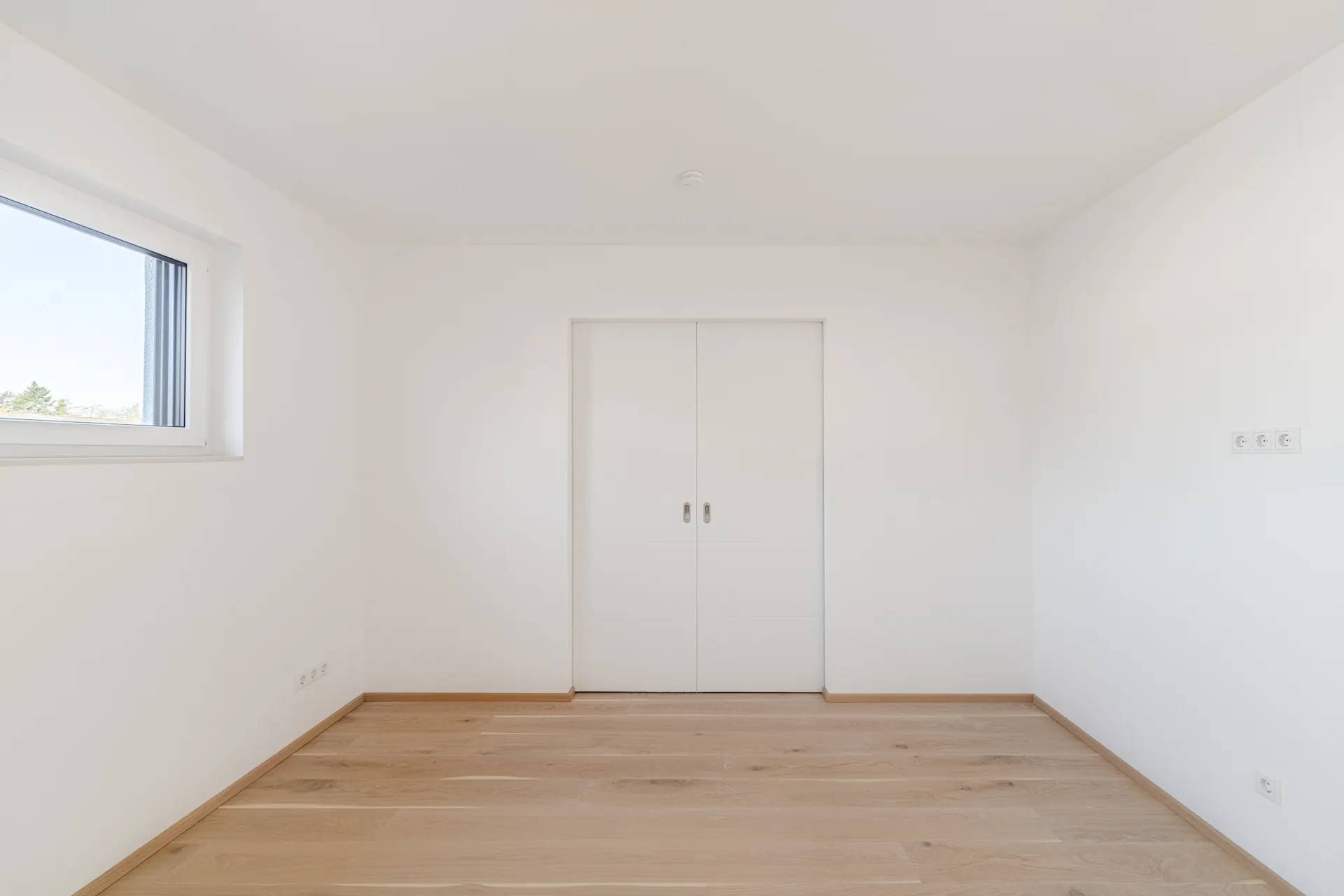 Leerer, heller Raum mit Holzfußboden, Fenster links und weißen Schiebetüren an der Wand.