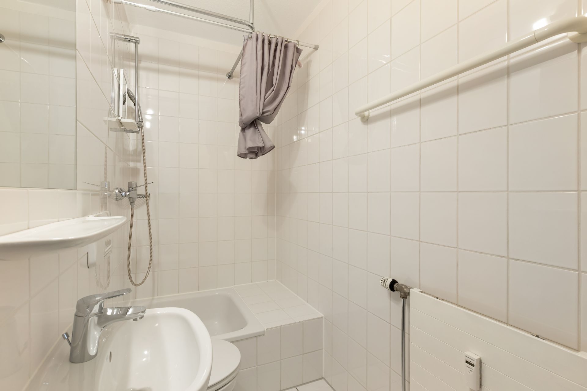 Kleines Badezimmer mit weiß gefliesten Wänden, Waschbecken, Dusche, Badewanne und Heizkörper.