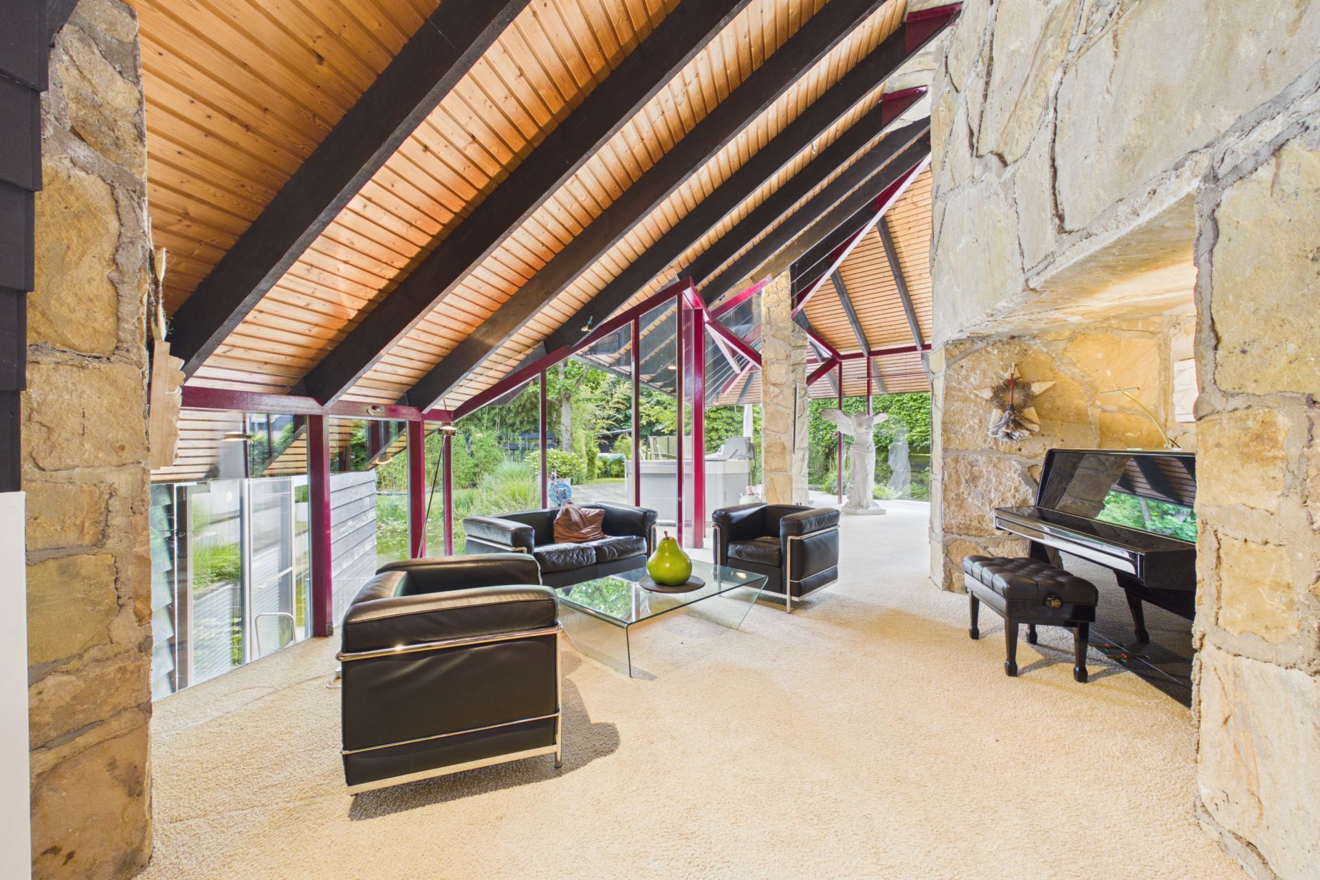 Modernes Wohnzimmer mit großen Fenstern, Ledersesseln, Glastisch und Klavier, sichtbare Holzbalkendecke.