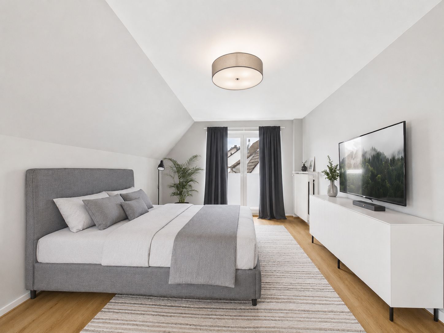 Modernes Schlafzimmer mit grauem Bett, Teppich, TV und bodentiefen Fenstern mit dunklen Vorhängen.