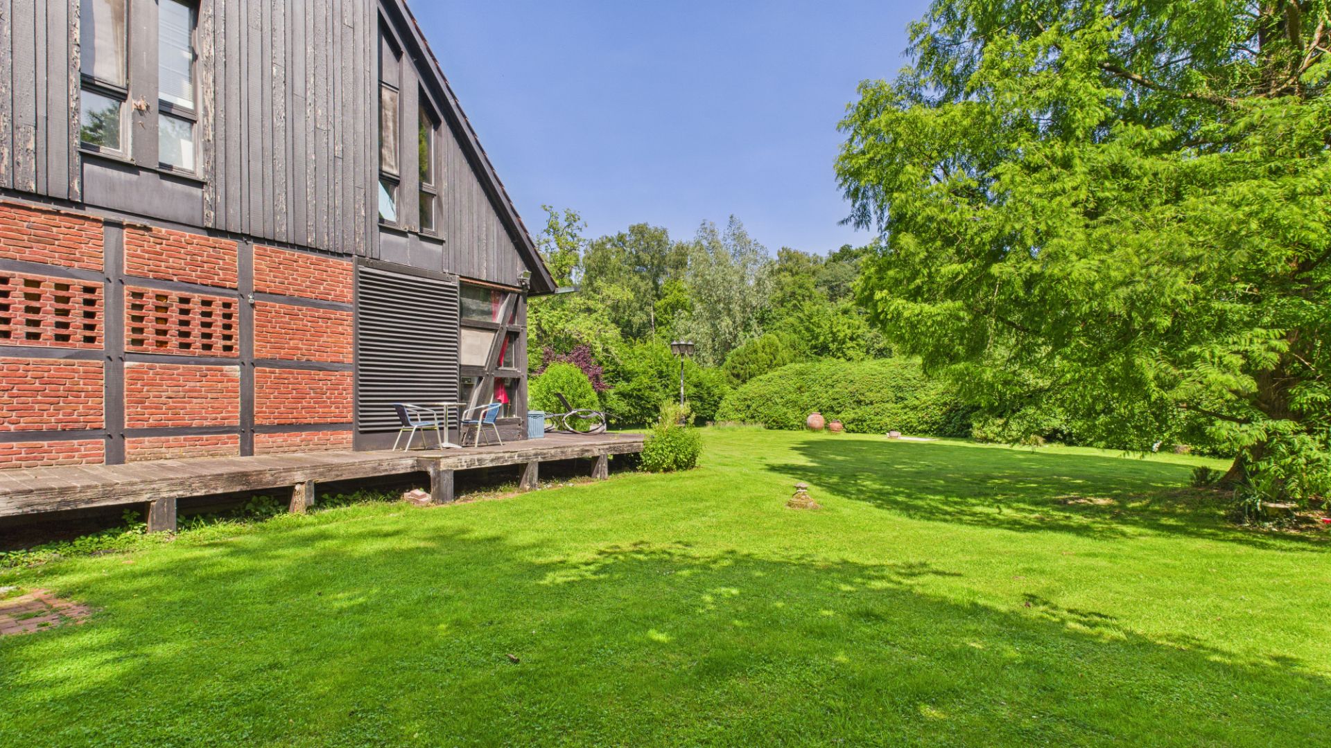 Haus mit Holz- und Ziegelfassade, davor Terrasse und großer grüner Garten mit Bäumen.