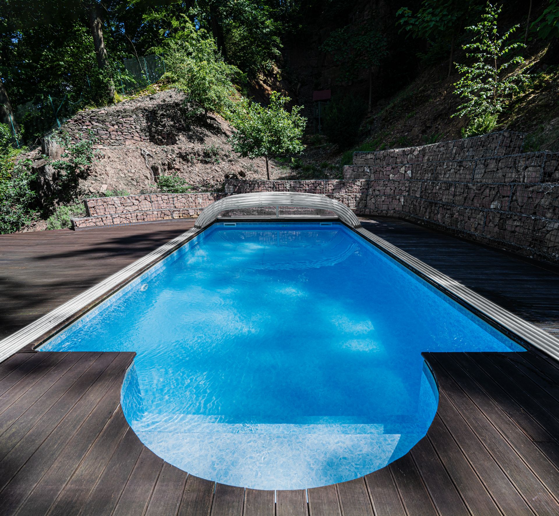 Blauer Swimmingpool im Garten mit Holzterrasse und steinernen Mauern im Hintergrund.