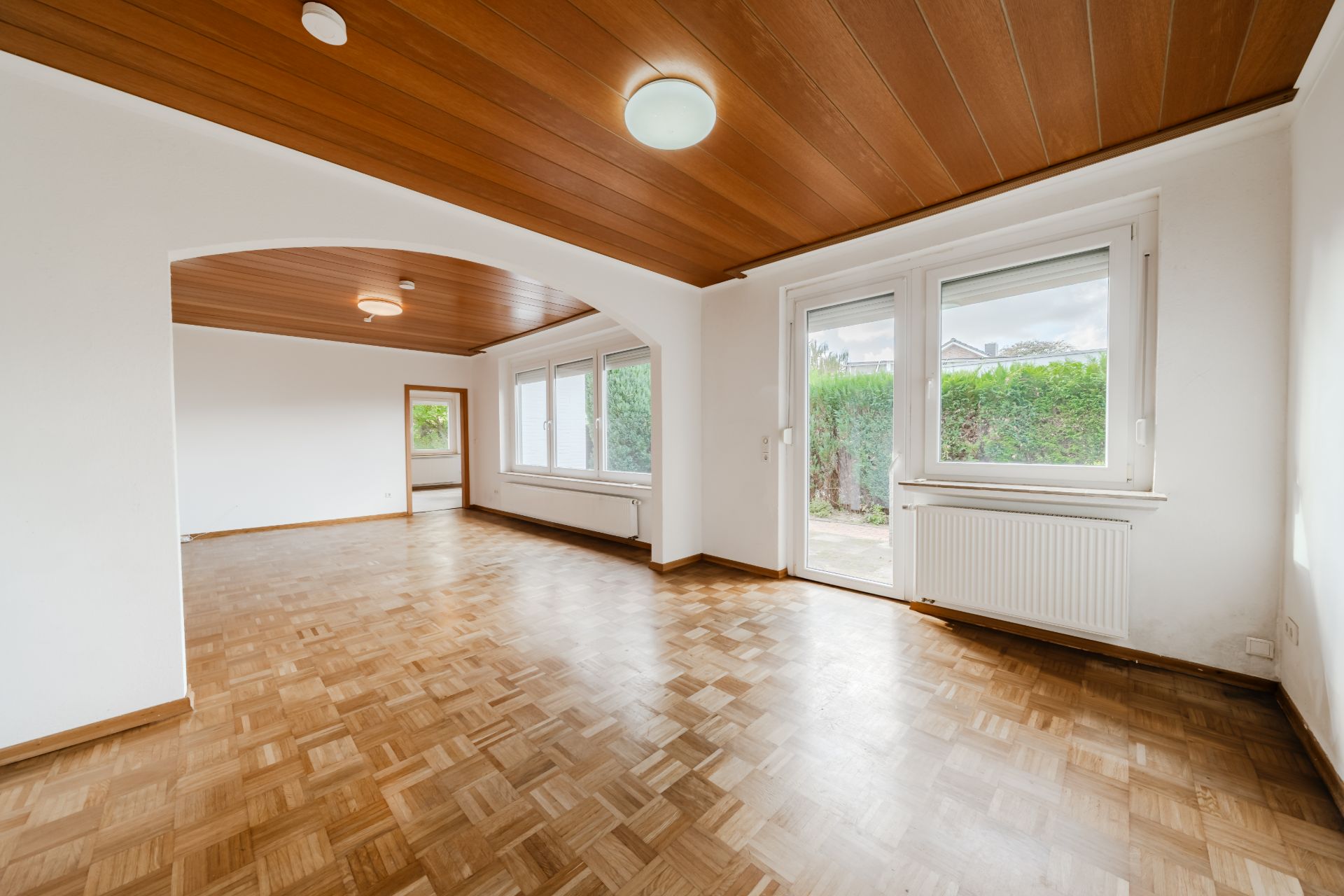 Heller, leerer Wohnraum mit Parkettboden, Holzdecke und großen Fenstern zum Garten.