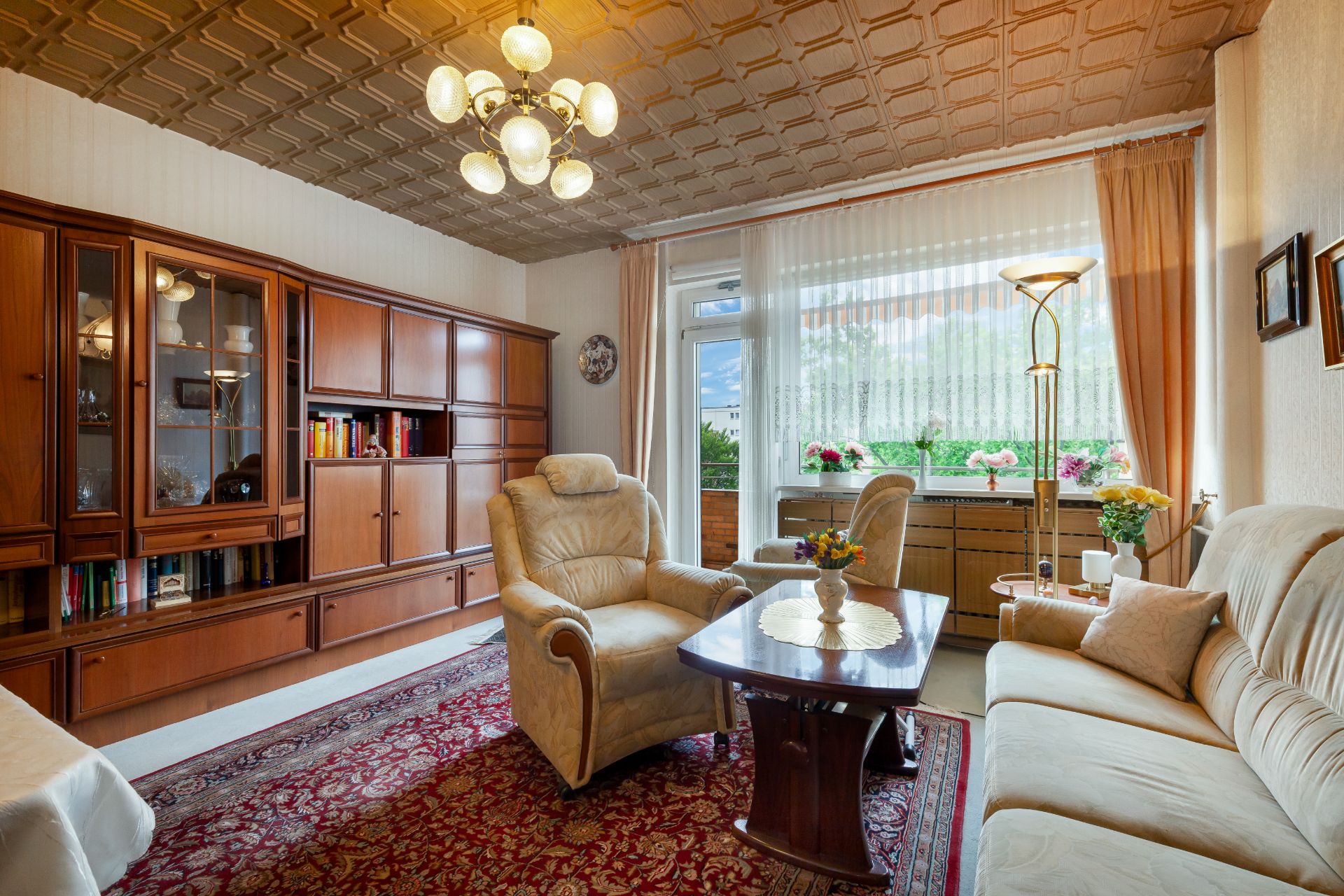 Wohnzimmer mit Teppich, Sofagarnitur, Holzschrankwand und großen Fenstern mit Vorhängen.