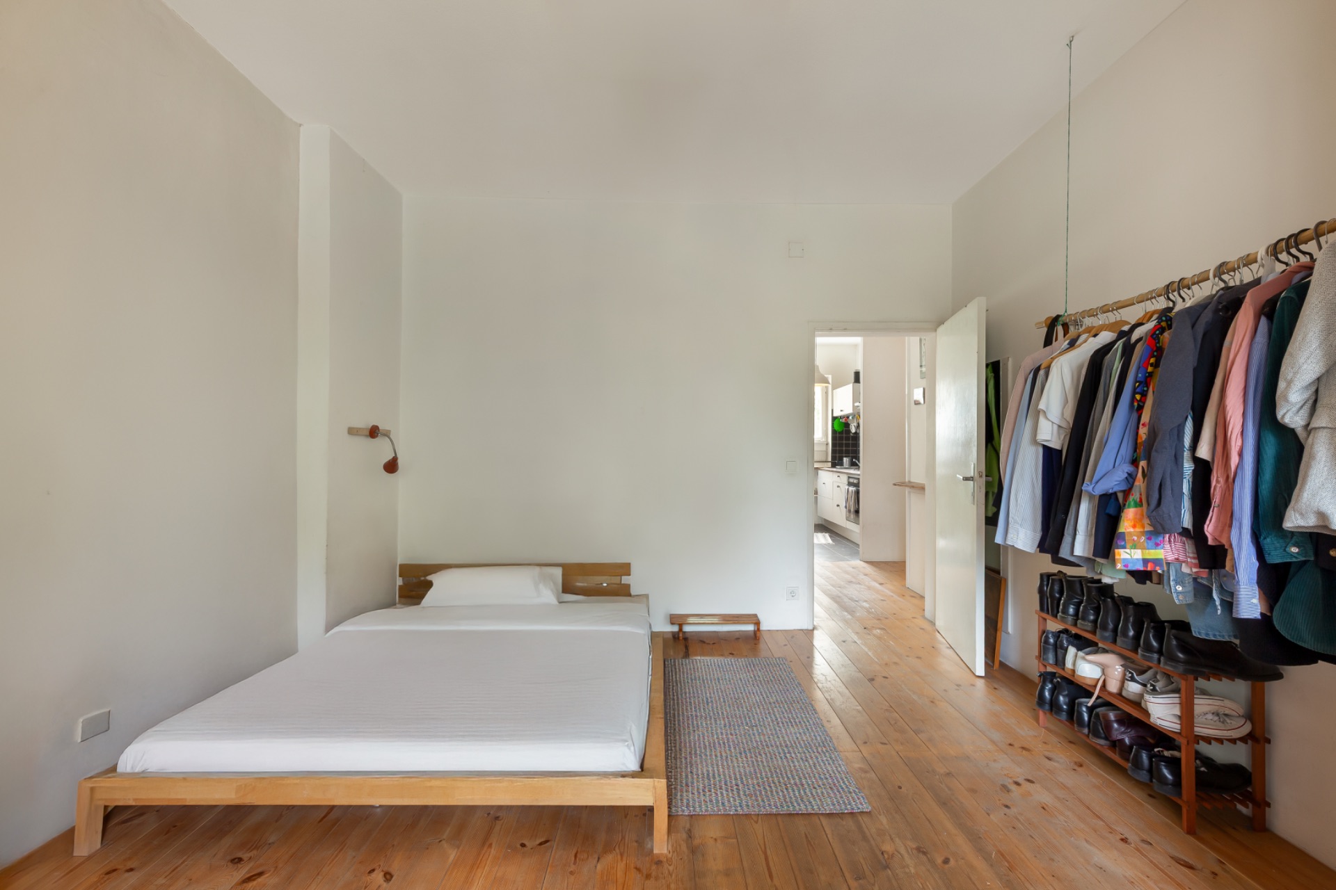 Schlafzimmer mit Holzfußboden, niedrigem Bett, Teppich und offener Kleiderstange mit Kleidung.