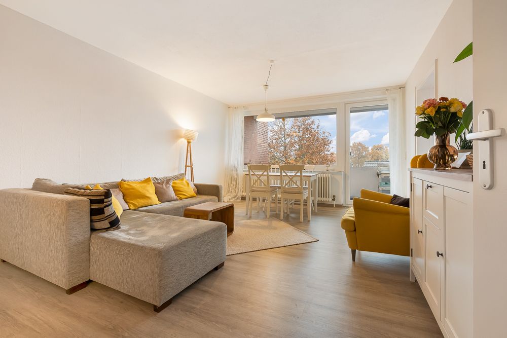 Helles Wohnzimmer mit Sofa, gelbem Sessel, Esstisch, großen Fenstern und Sideboard mit Blumen.