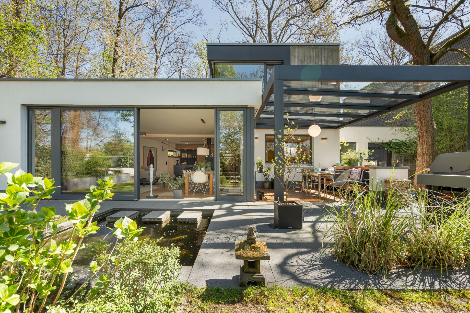 Moderne Terrasse mit Glasdach, Teich und Gartenmöbeln vor einem hellen Wohnhaus.