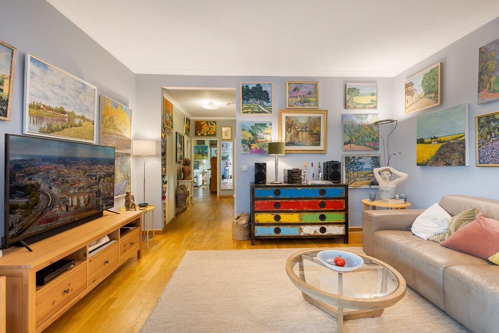 Wohnzimmer mit Ledersofa, TV, buntem Sideboard und vielen Gemälden an den Wänden.