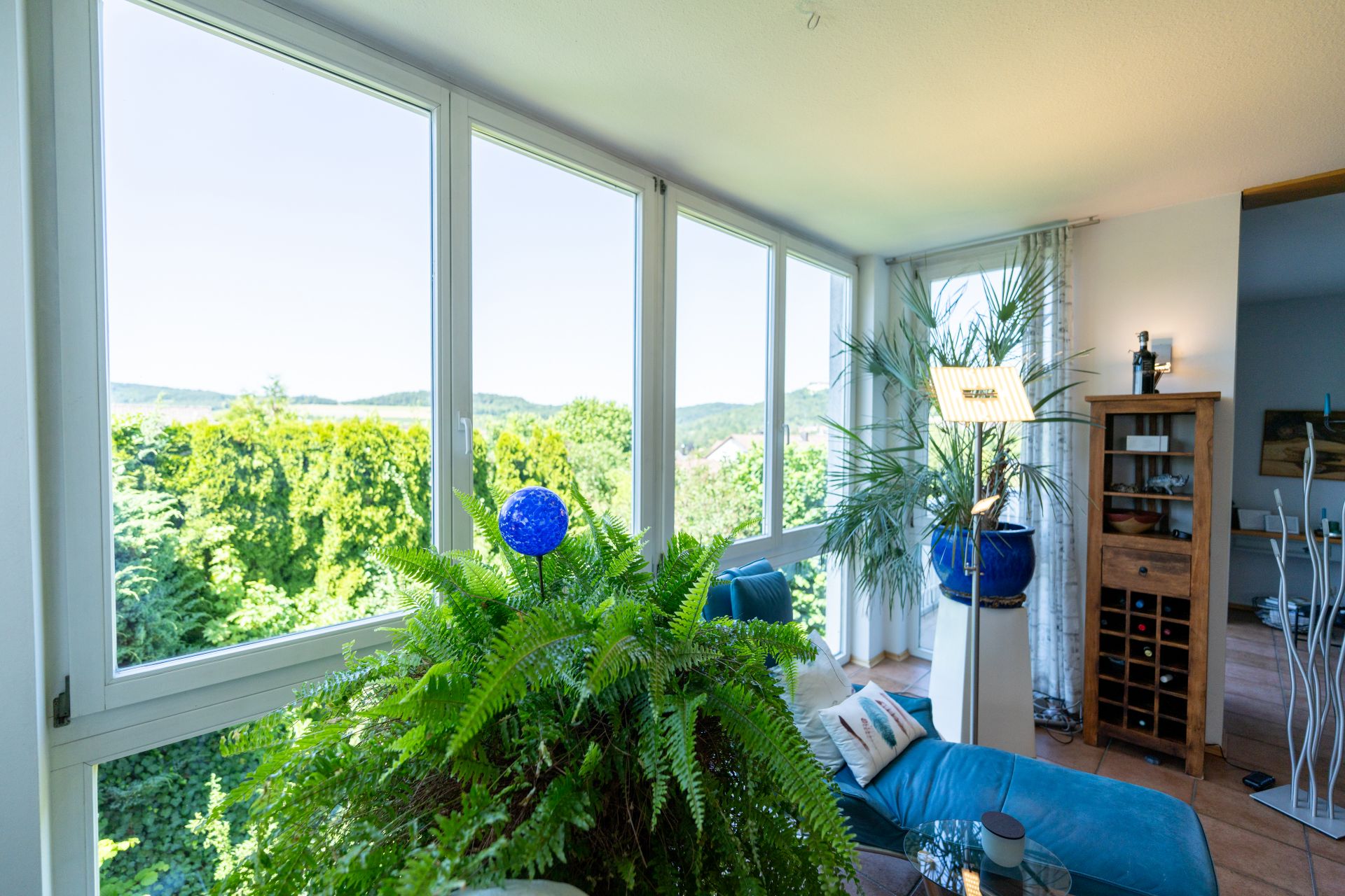 Helles Wohnzimmer mit großen Fenstern, vielen Pflanzen und Blick ins Grüne.