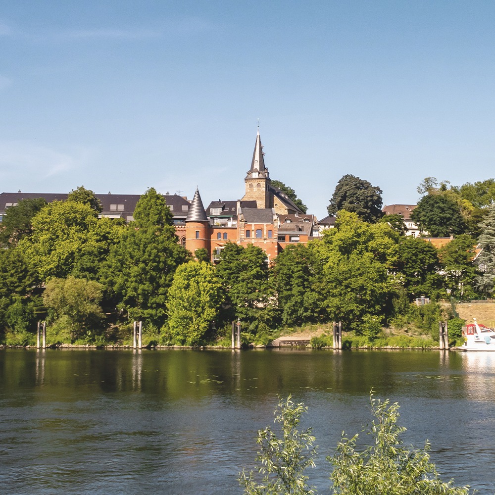 Backsteingebäude mit Turm hinter Bäumen, davor ein Fluss bei Tageslicht.