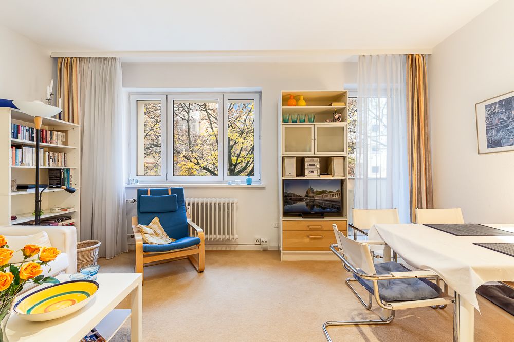 Heller Wohnraum mit Sessel, Esstisch, Regal und großen Fenstern mit Blick auf Bäume.