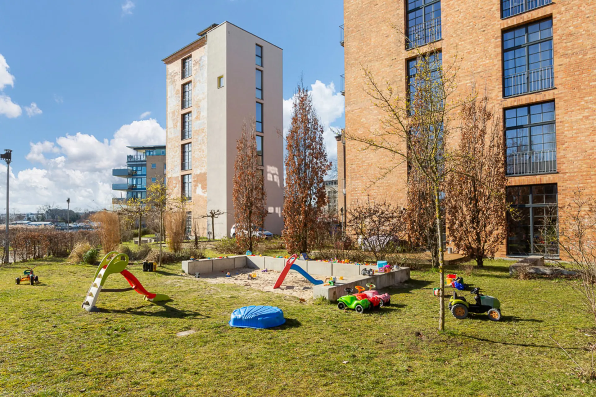 Grüner Hof mit Sandkasten, Kinderrutsche und Spielzeug, umgeben von Wohngebäuden.