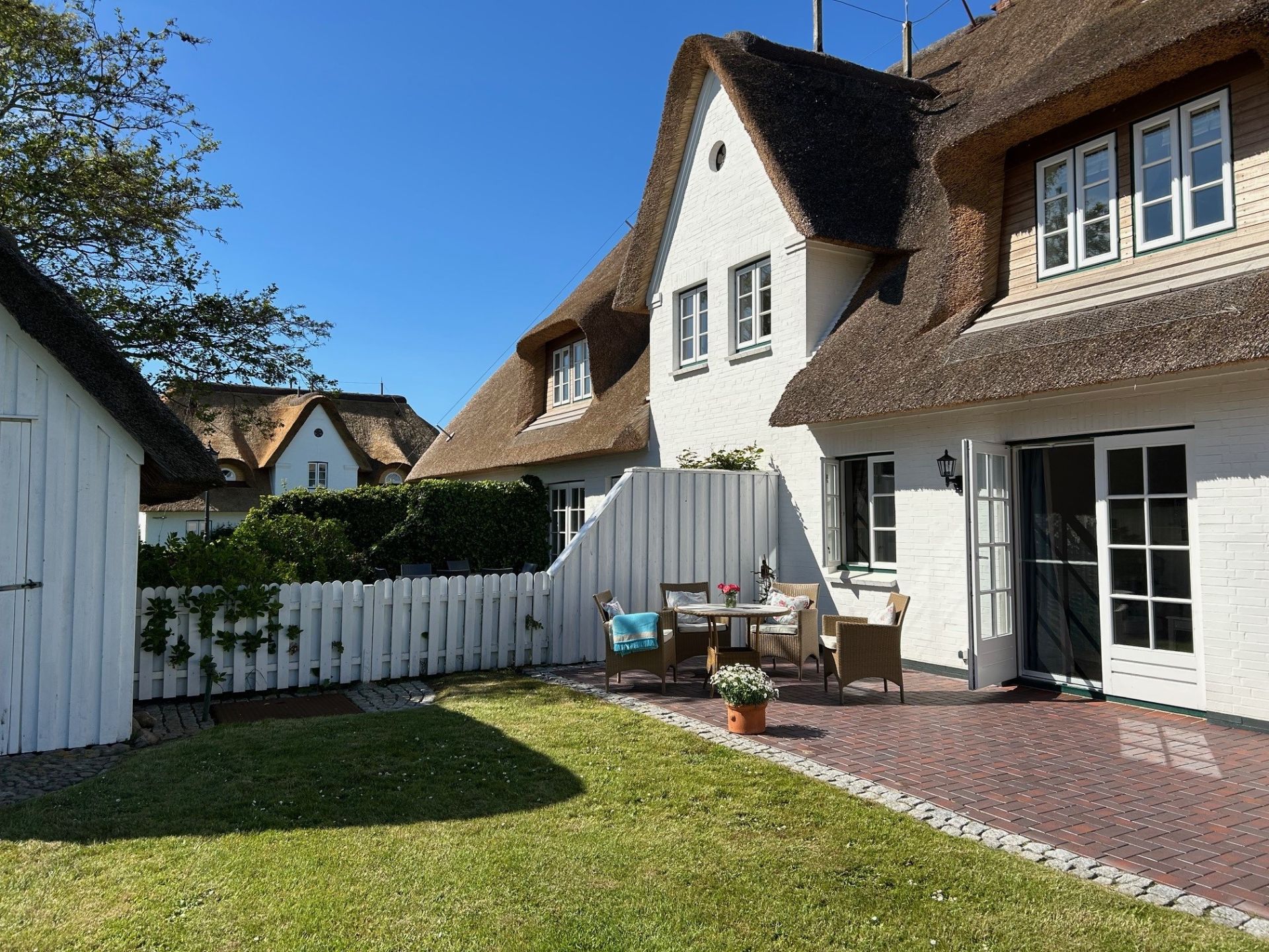 Garten mit Terrasse, Sitzgruppe, weißem Zaun und Reetdachhaus bei Sonnenschein.