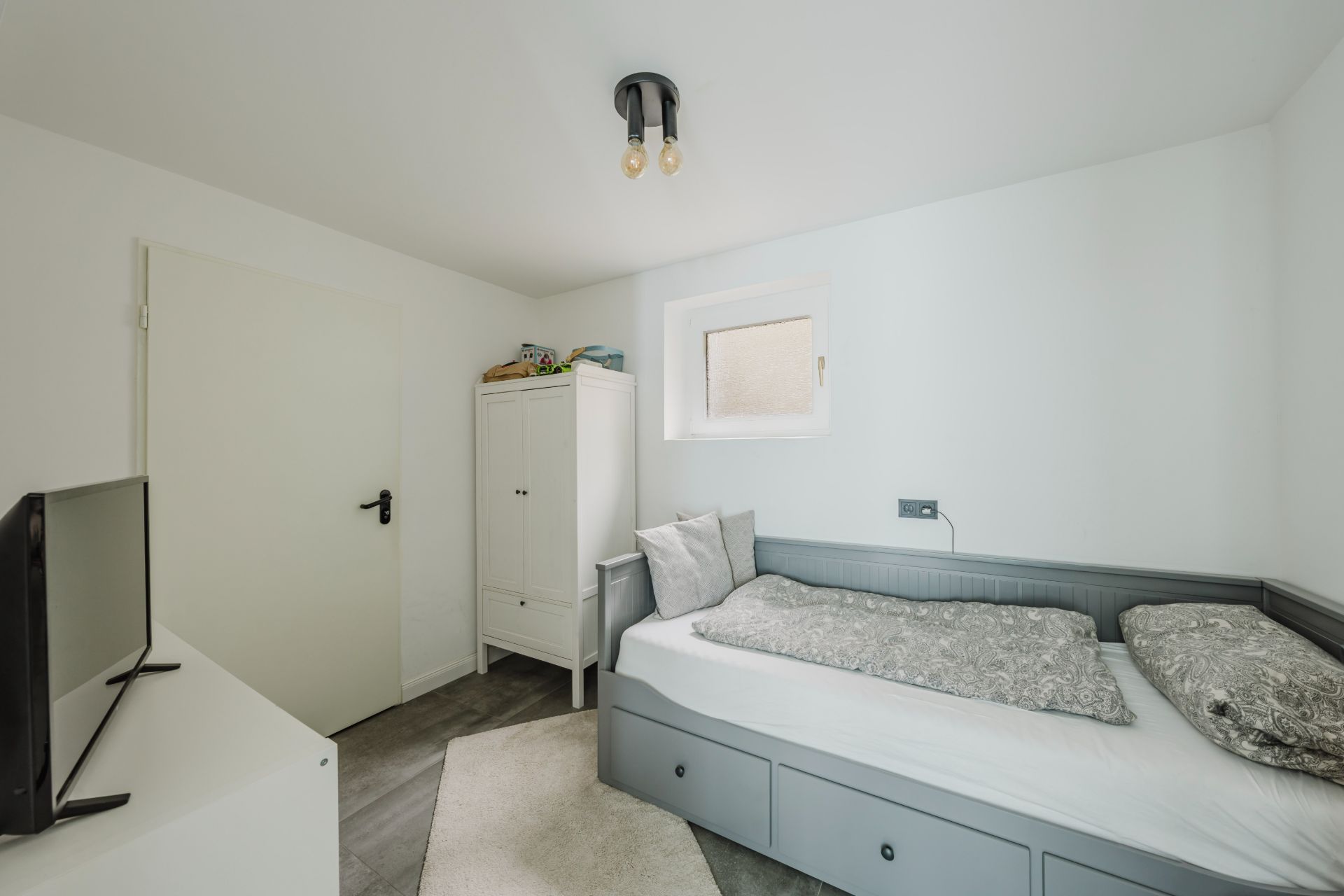 Kleines Schlafzimmer mit Einzelbett, grauer Kommode, weißem Schrank und Fernseher.
