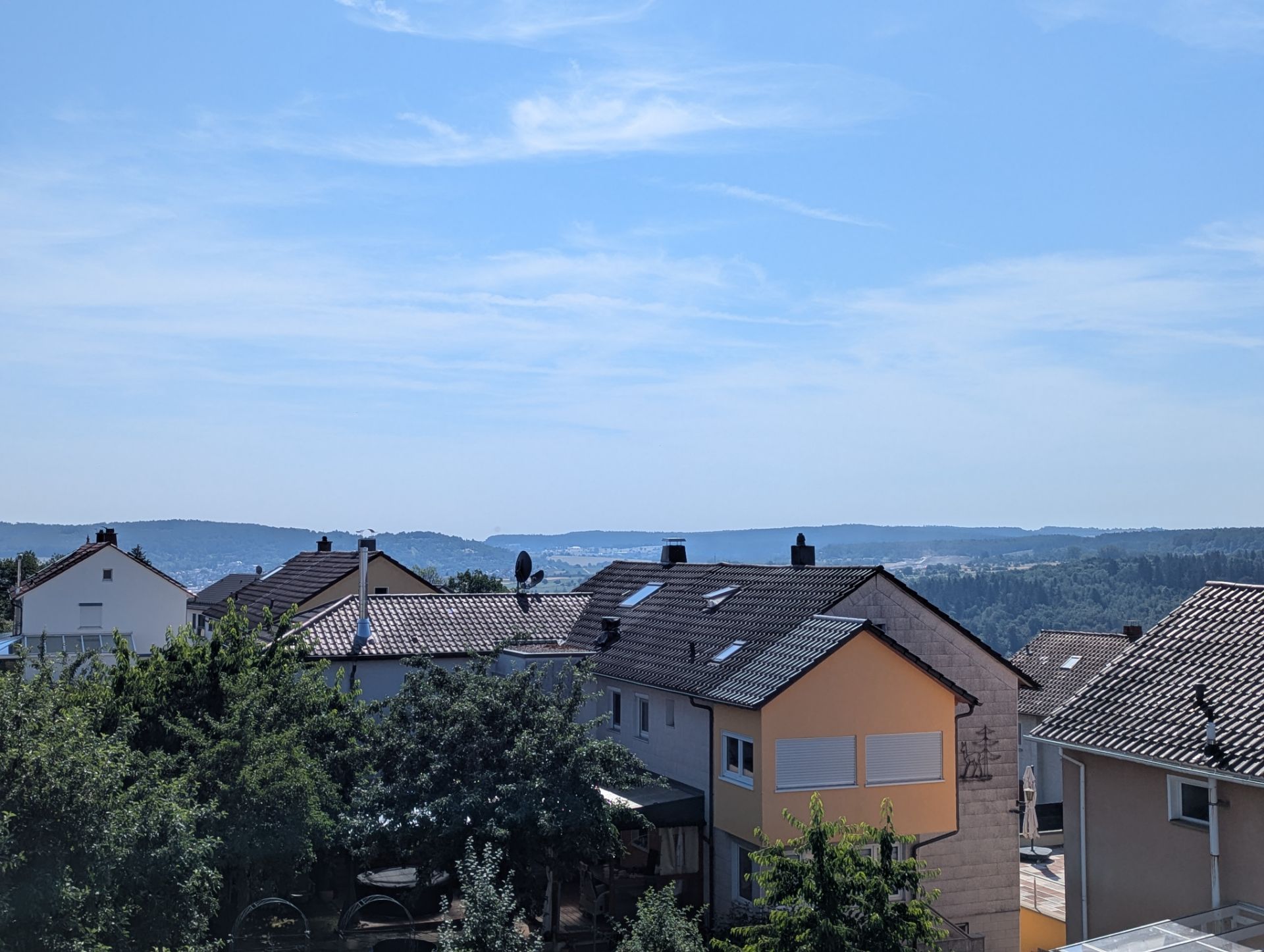 Blick auf mehrere Wohnhäuser mit Gärten unter blauem Himmel und Hügeln im Hintergrund.