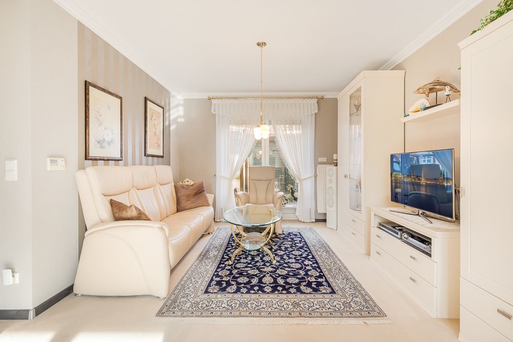 Helles Wohnzimmer mit beiger Couch, Teppich, Glastisch, Sessel, TV und großen Fenstern mit Vorhängen.