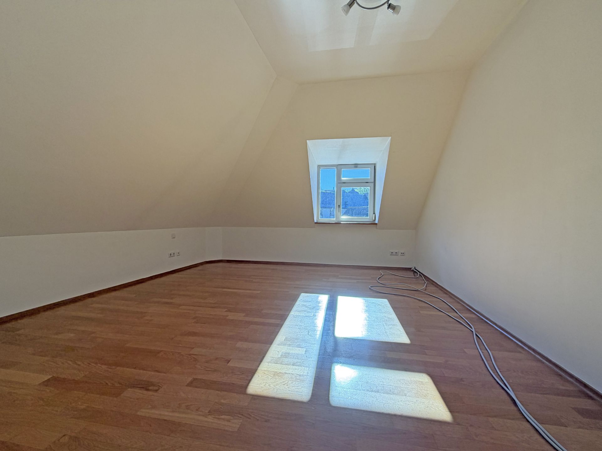 Leeres Dachzimmer mit Holzfußboden, Fenster und sichtbaren Kabeln auf dem Boden.
