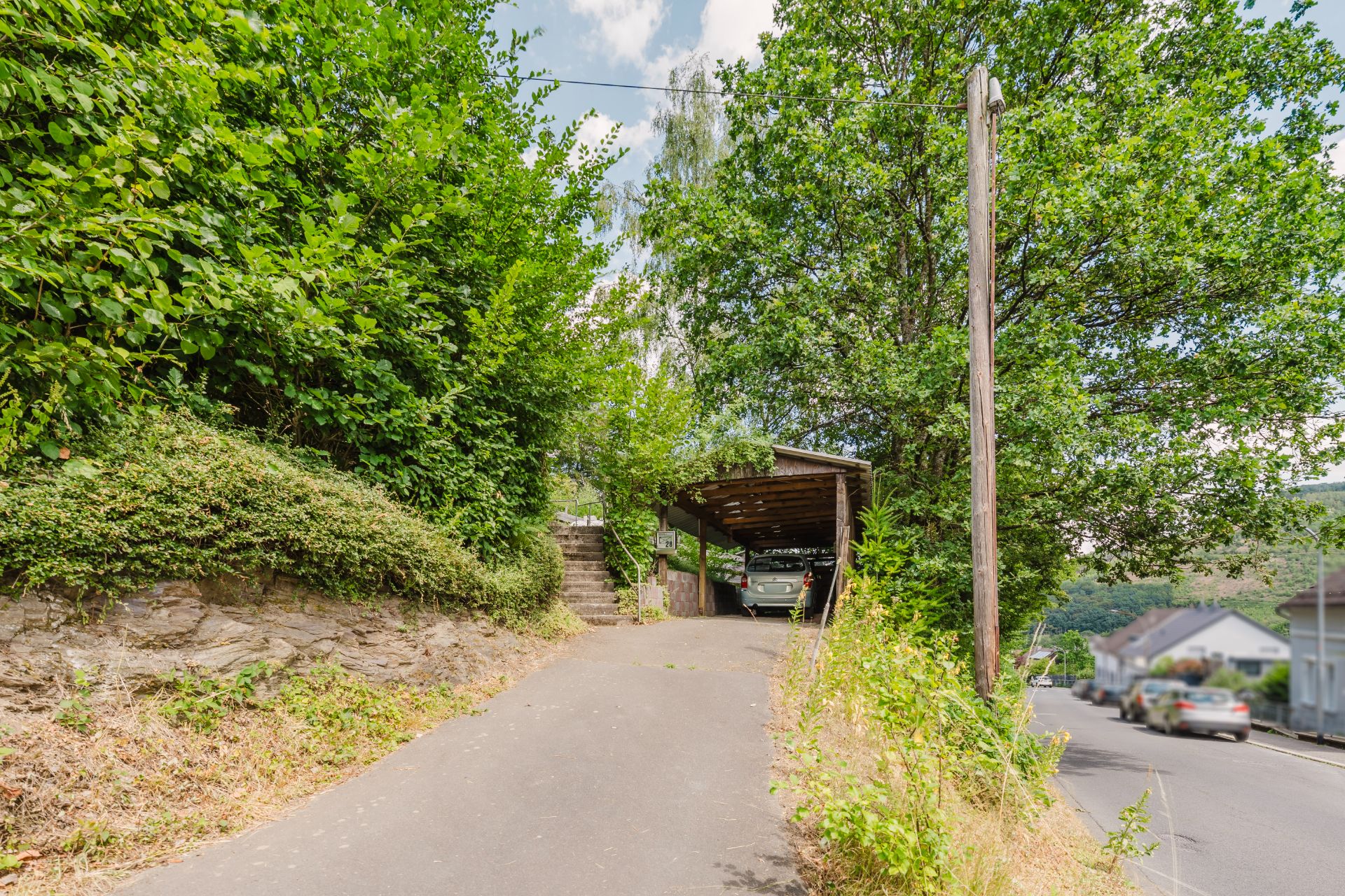 Auffahrt mit Carport, Auto darunter, umgeben von Bäumen und einer Straße rechts.