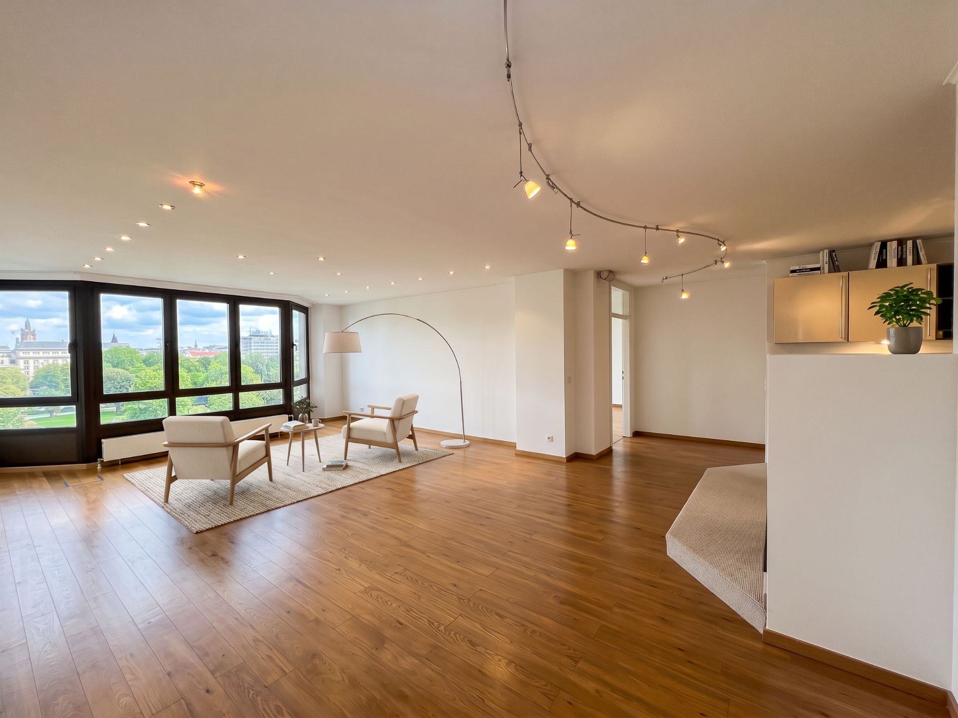 Großes, helles Wohnzimmer mit Holzfußboden, Sitzbereich und großen Fenstern mit Blick ins Grüne.
