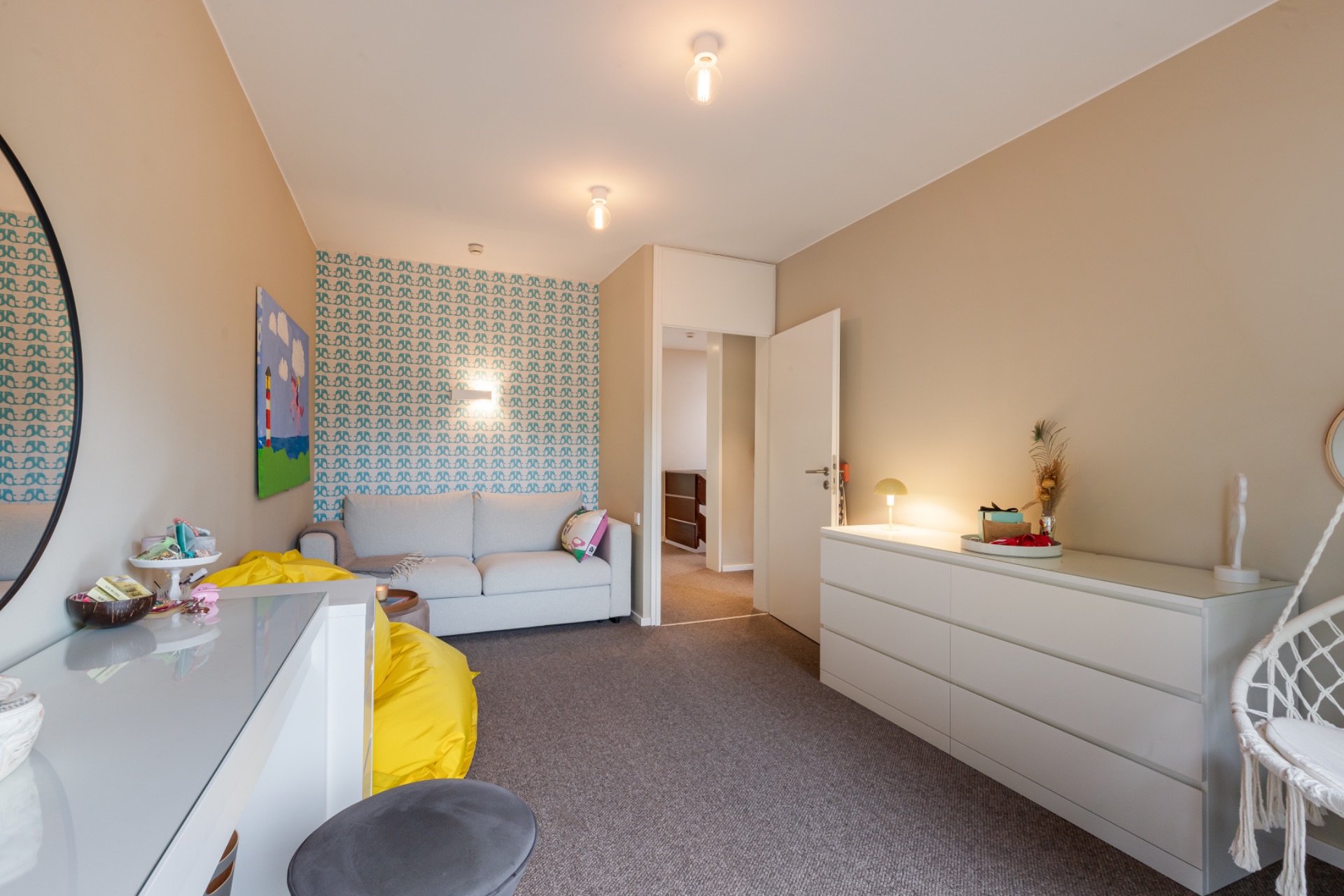 Helles Zimmer mit Sofa, gelbem Sitzsack, Kommode und gemusterter Tapete an einer Wand.