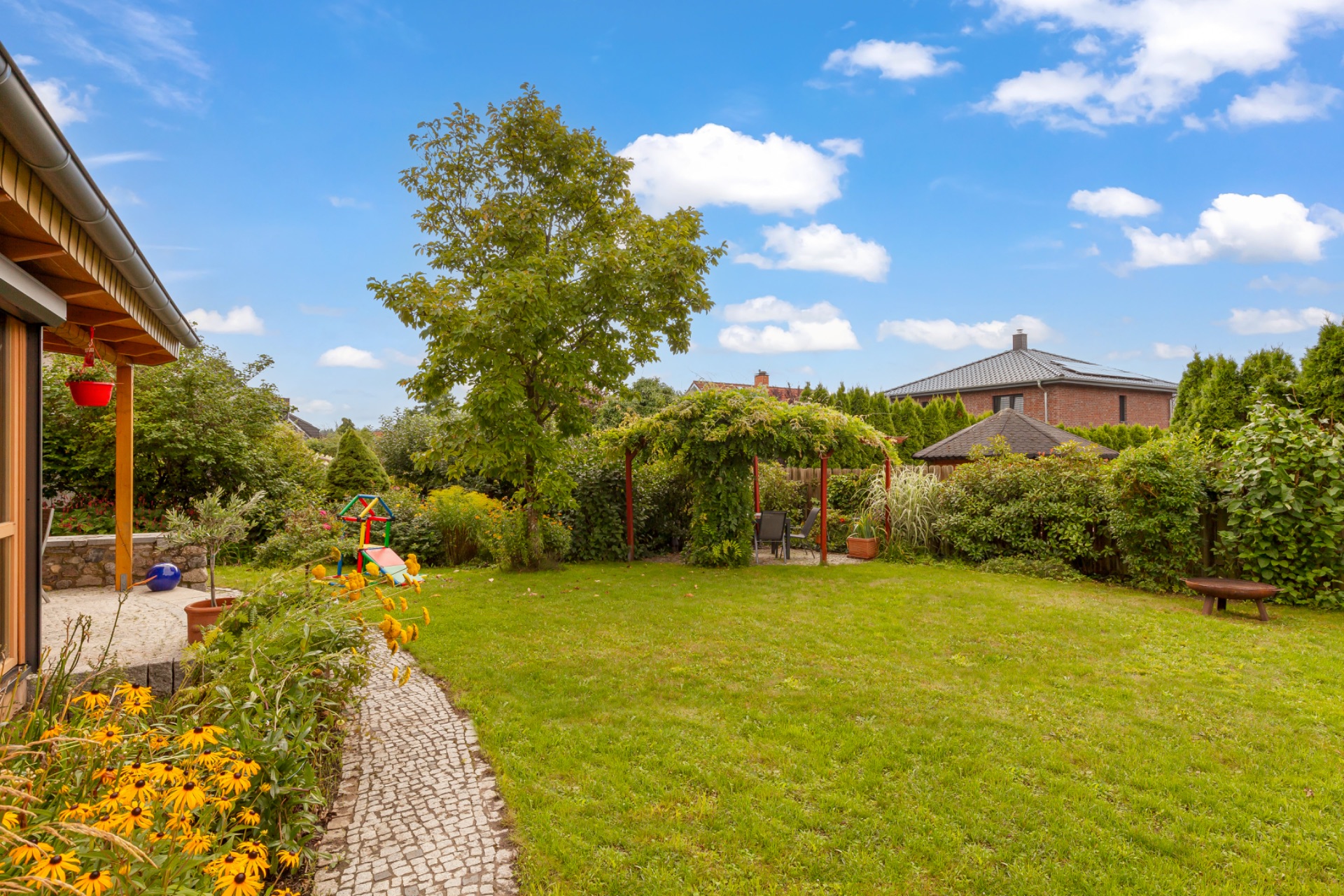 Garten mit Rasen, Baum, Sitzbereich unter Pergola und Blumen, Weg aus Pflastersteinen.