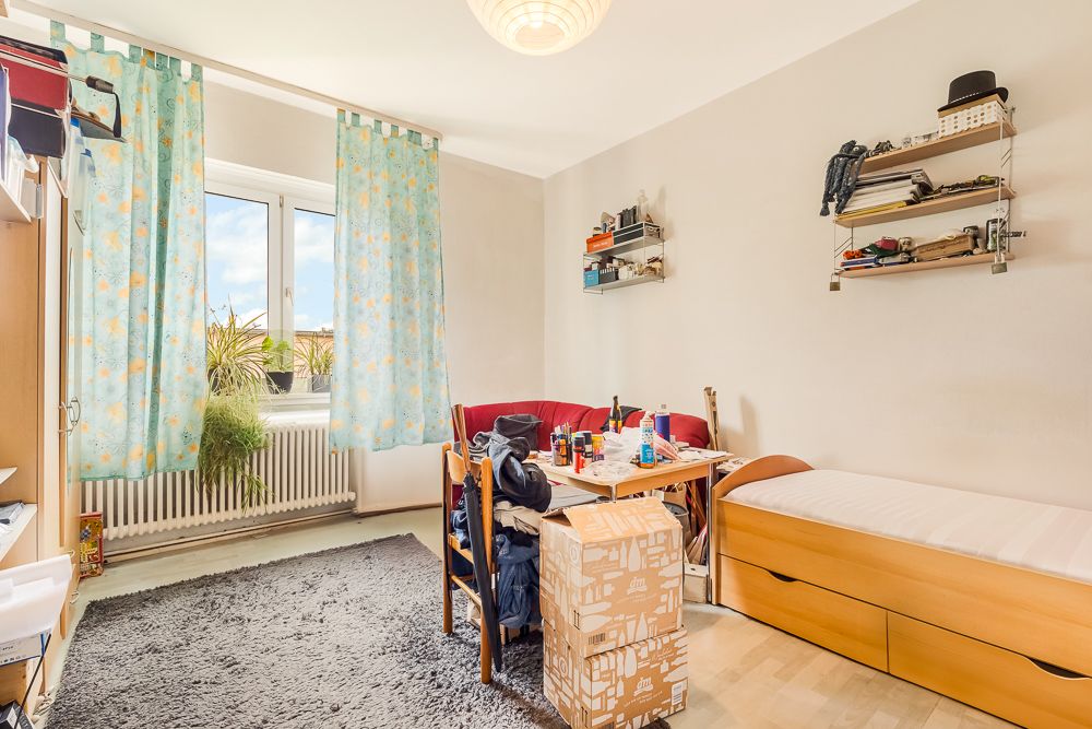 Helles Zimmer mit Einzelbett, Tisch, Teppich, Regal, Pflanzen und Karton auf dem Boden.