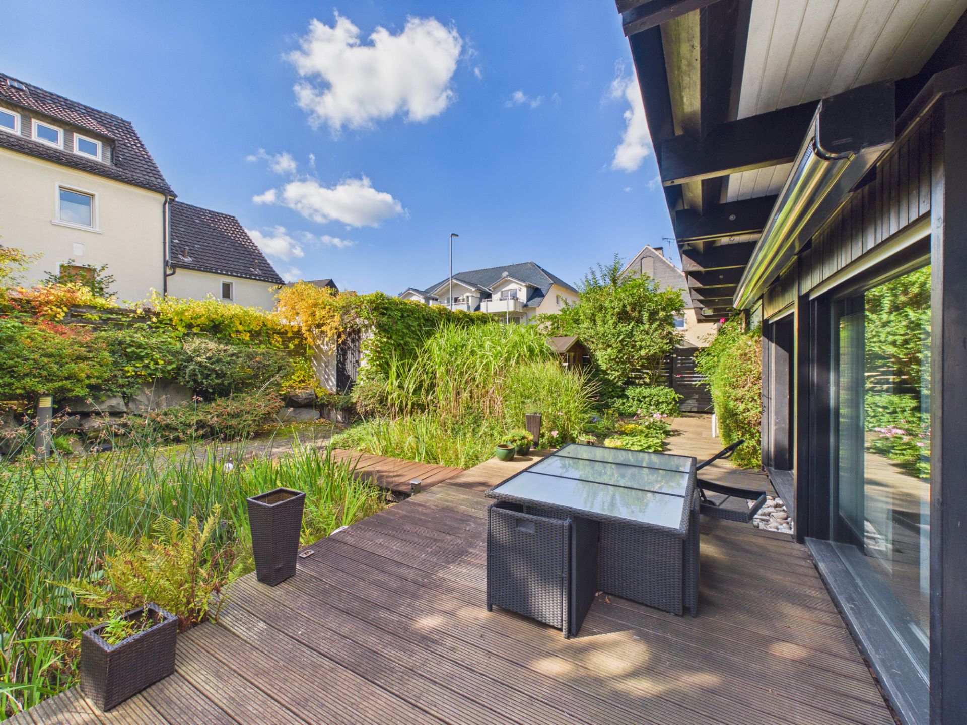 Holzterrasse mit Esstisch und Gartenblick, umgeben von Pflanzen und Nachbarhäusern.