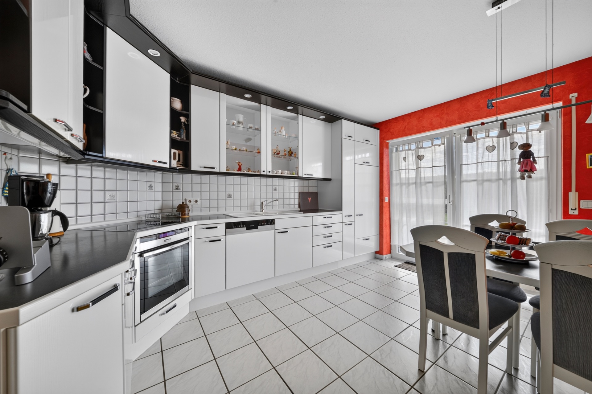 Moderne weiße Einbauküche mit Esstisch und großen Fenstern vor roter Wand.
