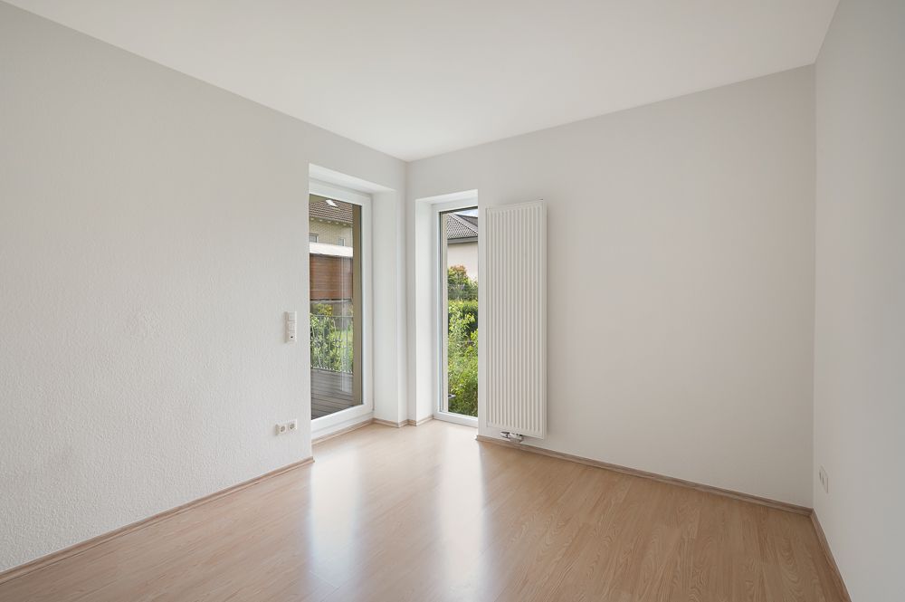 Leerer, heller Raum mit Parkettboden, großen Fenstern und Heizkörper an der Wand.