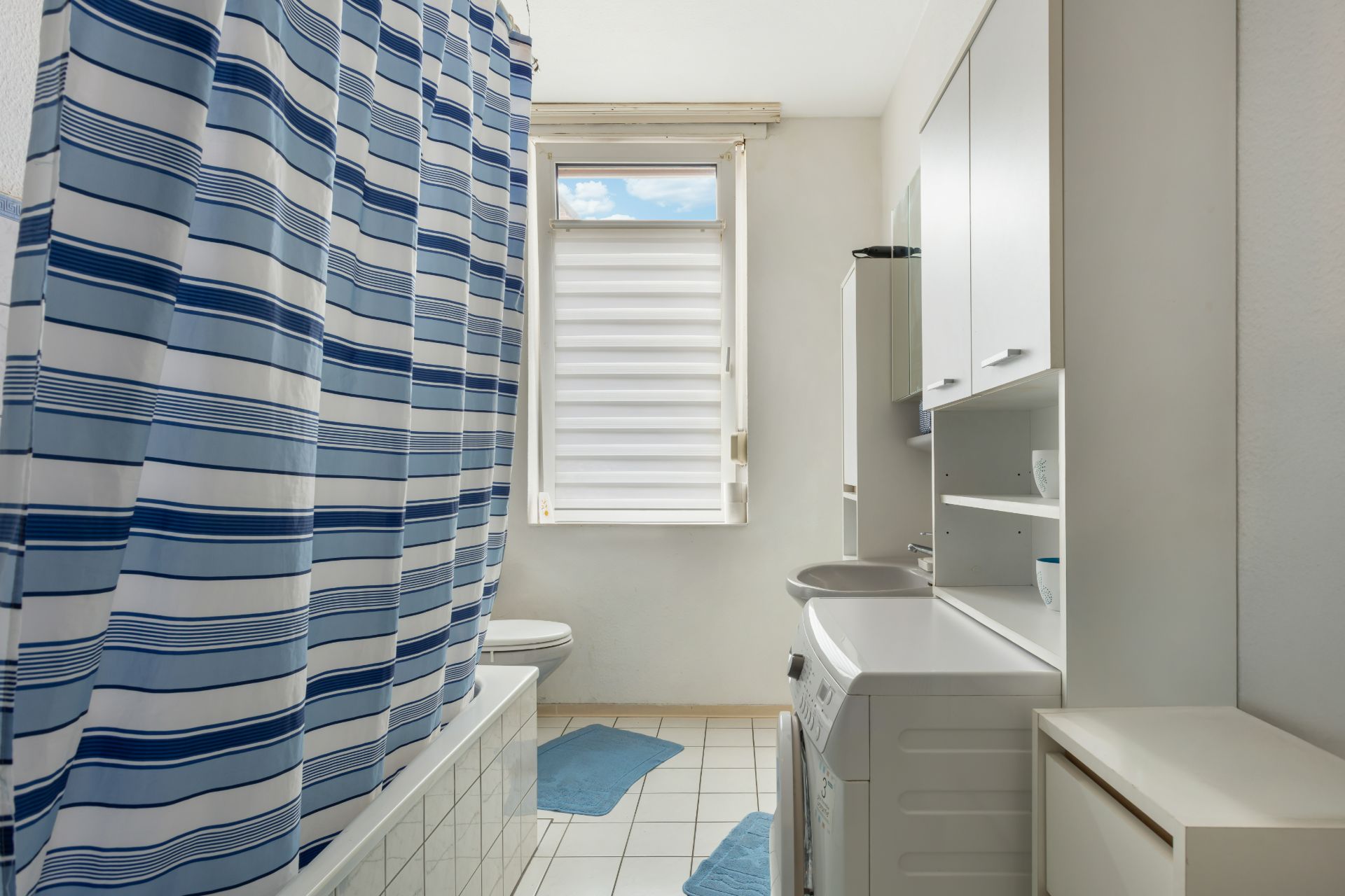 Helles Badezimmer mit Badewanne, blauem Duschvorhang, Waschmaschine und Fenster.