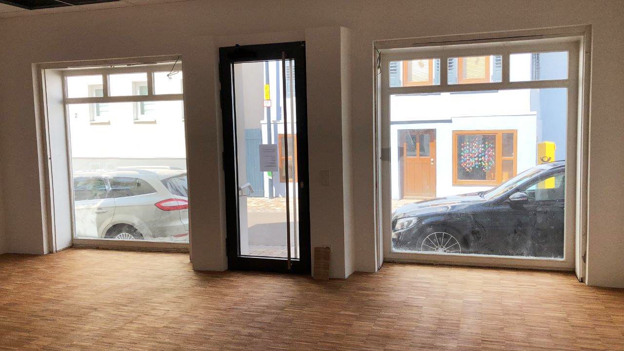 Heller Raum mit Parkettboden, zwei großen Fenstern und einer Glastür, Blick auf geparkte Autos.