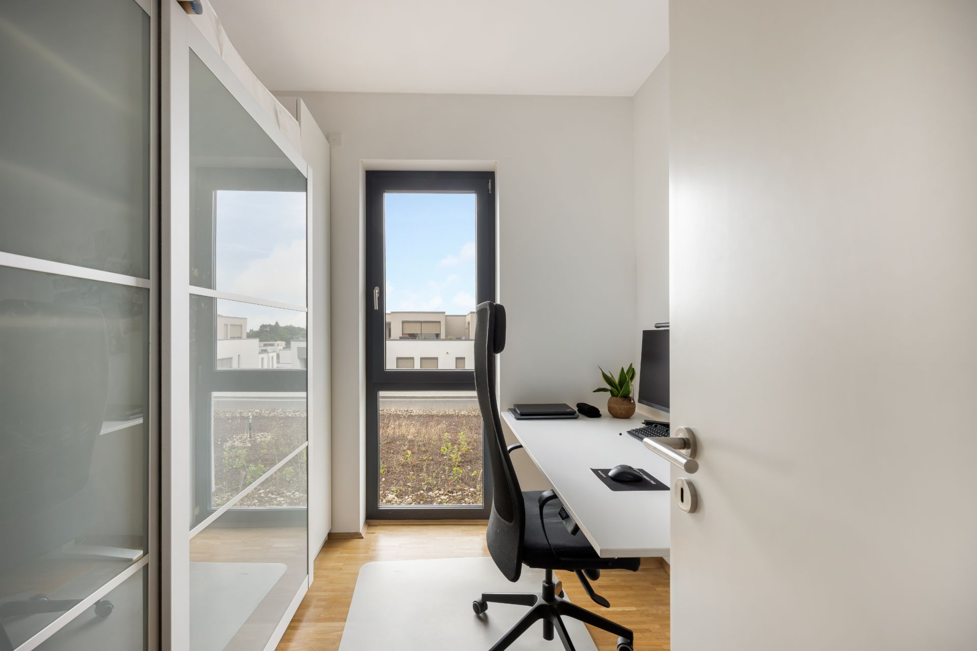 Helles Arbeitszimmer mit Schreibtisch, Stuhl, Computer und großem Fenster.