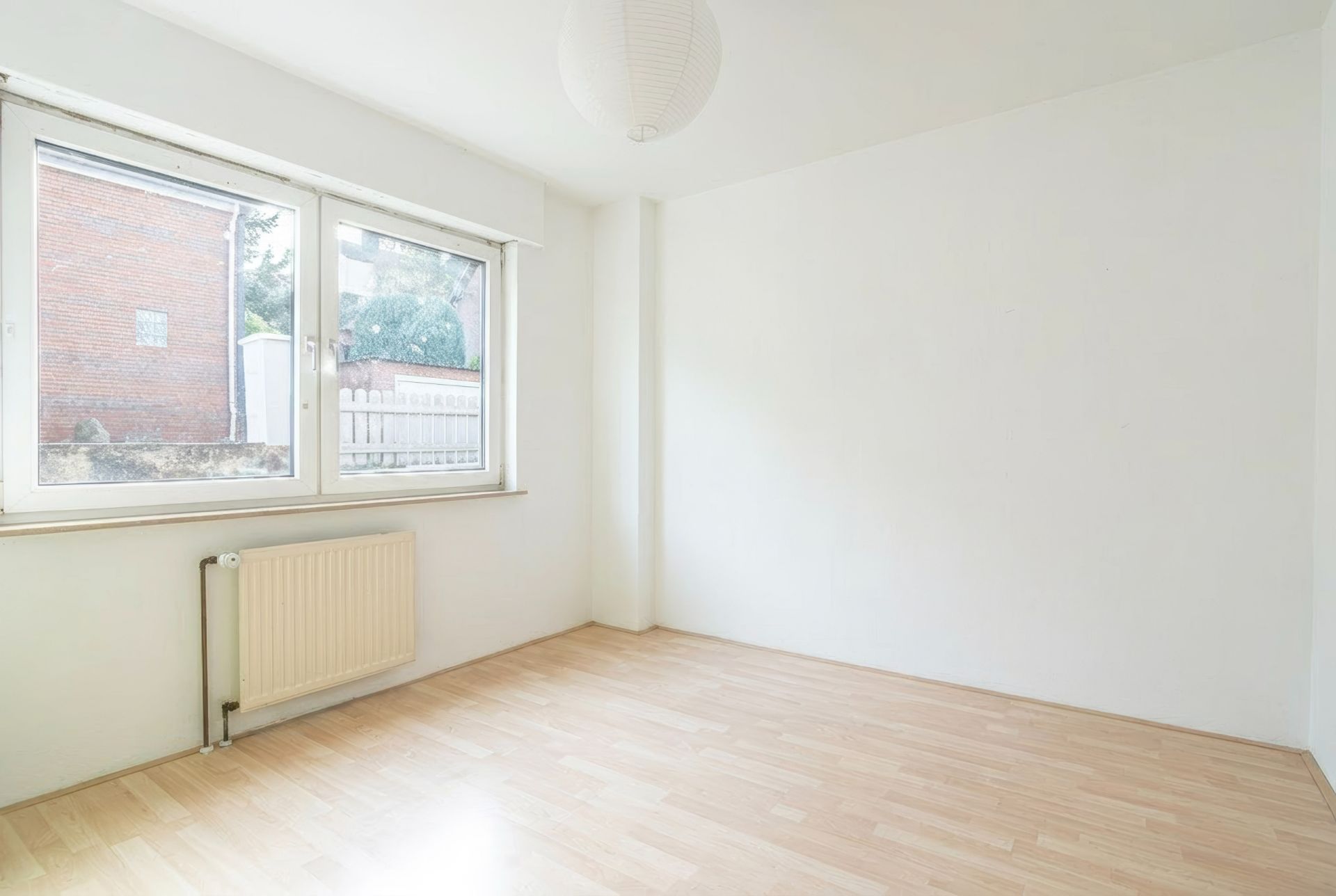 Leeres, helles Zimmer mit großem Fenster, Heizkörper und Holzfußboden.