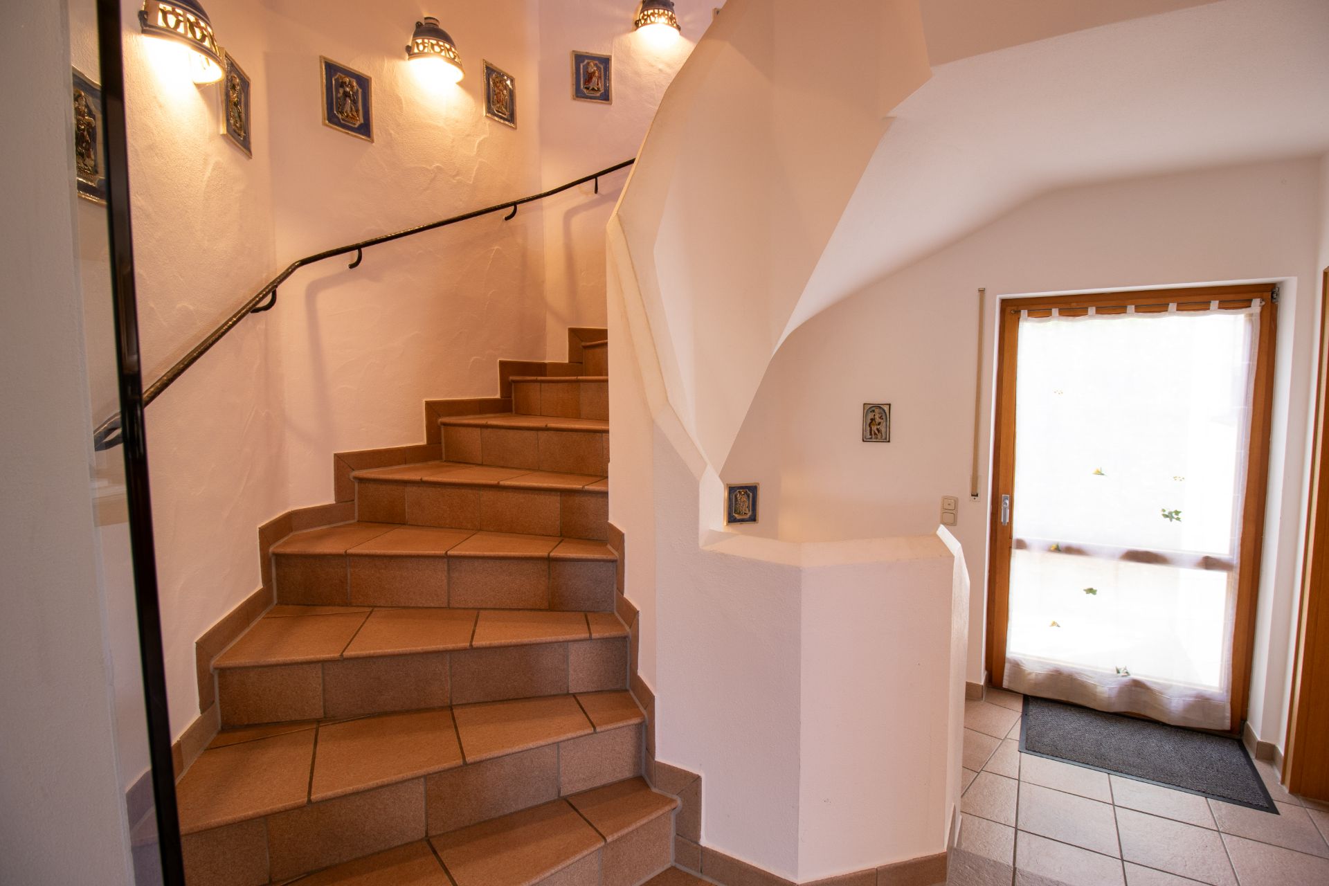 Treppenhaus mit braunen Fliesen, Handlauf und beleuchteten Wandlampen, daneben eine Glastür mit Vorhang.