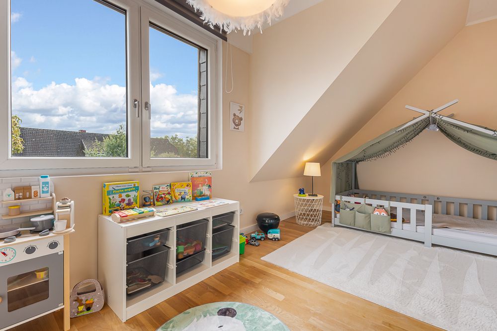 Helles Kinderzimmer mit großem Fenster, Spielküche, Bücherregal, Bett und Lampe.