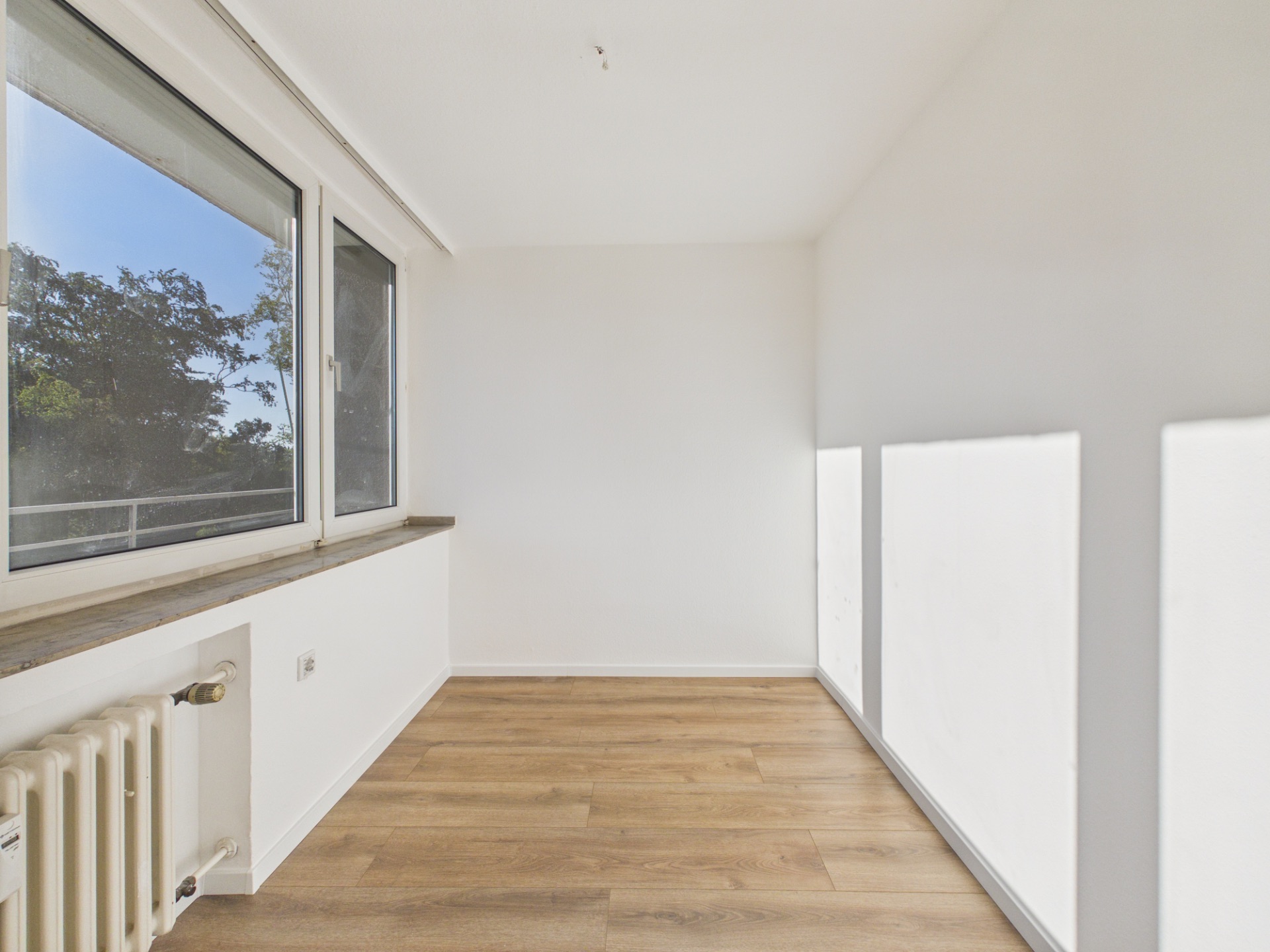 Heller, leerer Raum mit großem Fenster, Holzboden und Heizkörper.