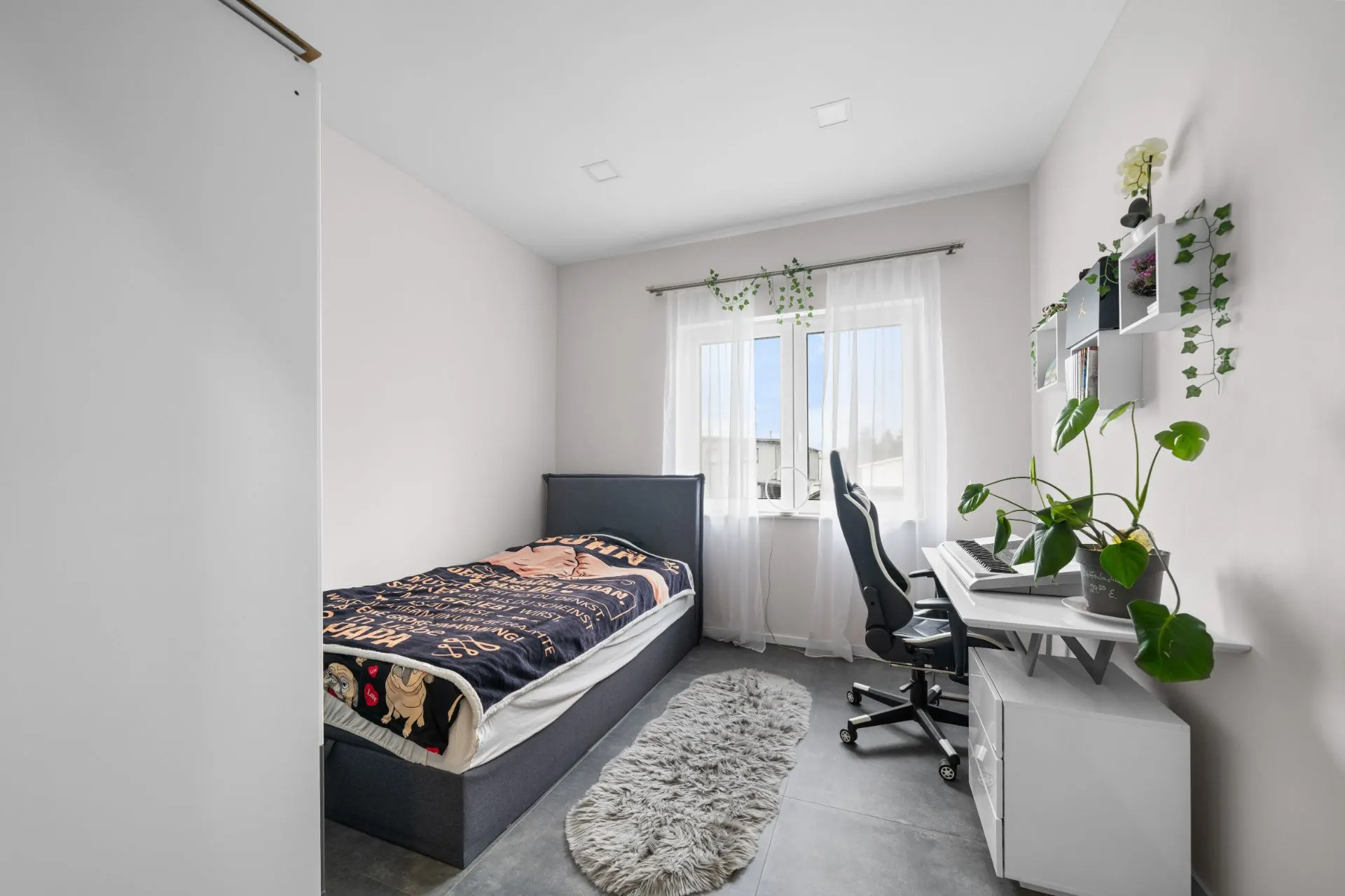 Helles Schlafzimmer mit Einzelbett, Schreibtisch, Bürostuhl und großen Pflanzen am Fenster.