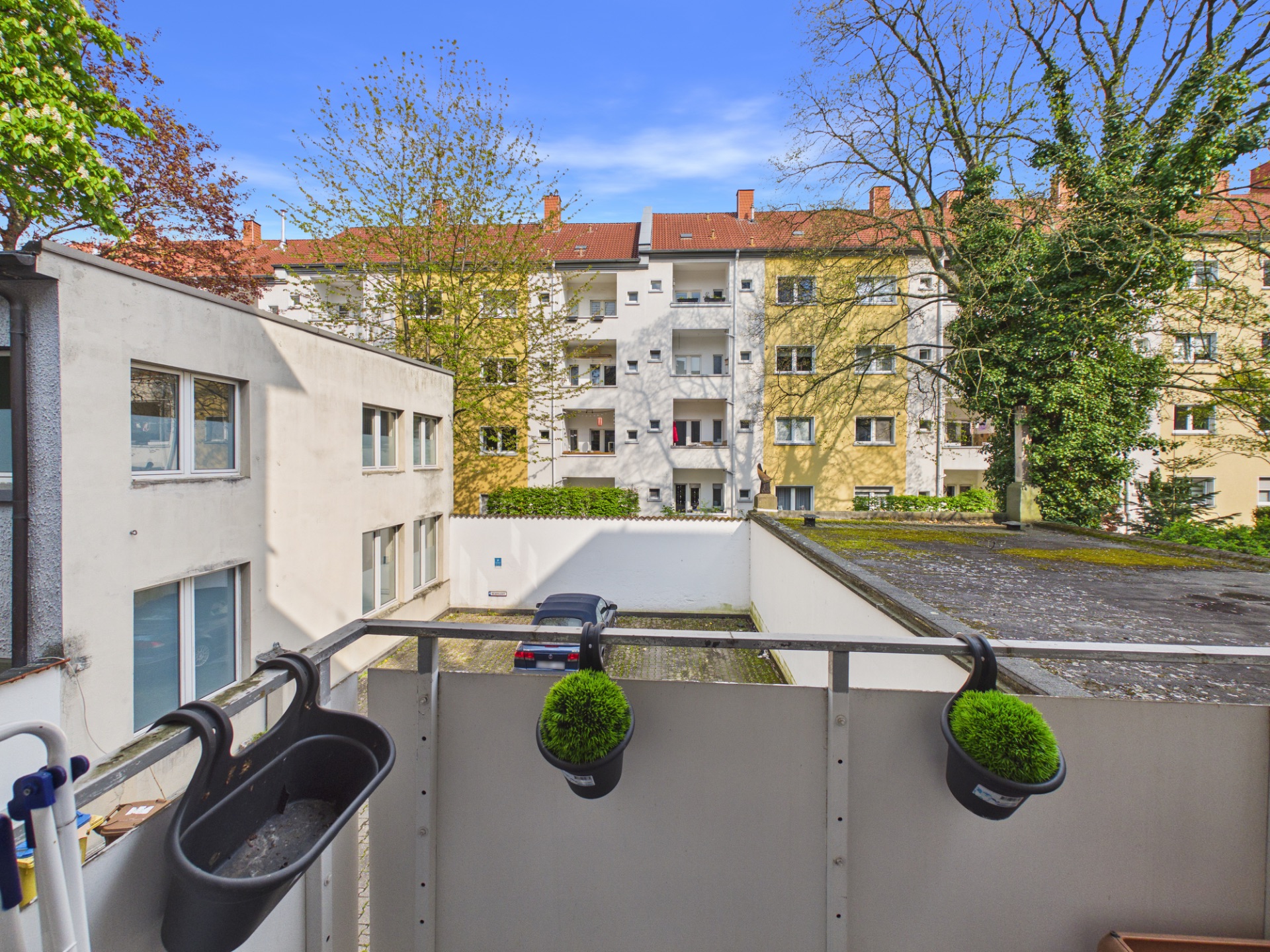 Blick von einem Balkon auf einen Innenhof mit geparktem Auto und Wohnhäusern im Hintergrund.
