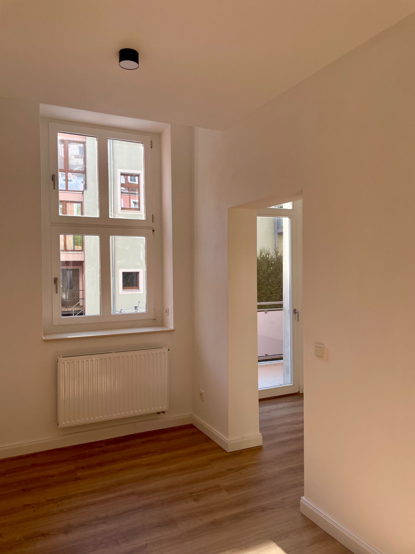 Heller Raum mit Holzfußboden, Fenster, Heizkörper und Zugang zu einem Balkon.