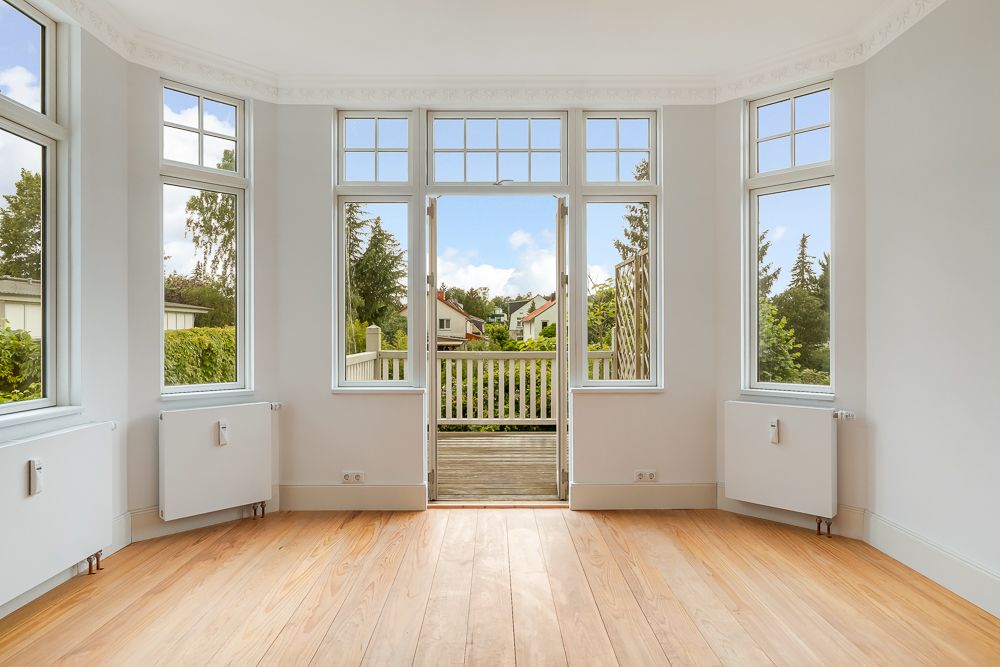 Heller Raum mit Holzboden, großen Fenstern und Tür zu einem Balkon mit Gartenblick.